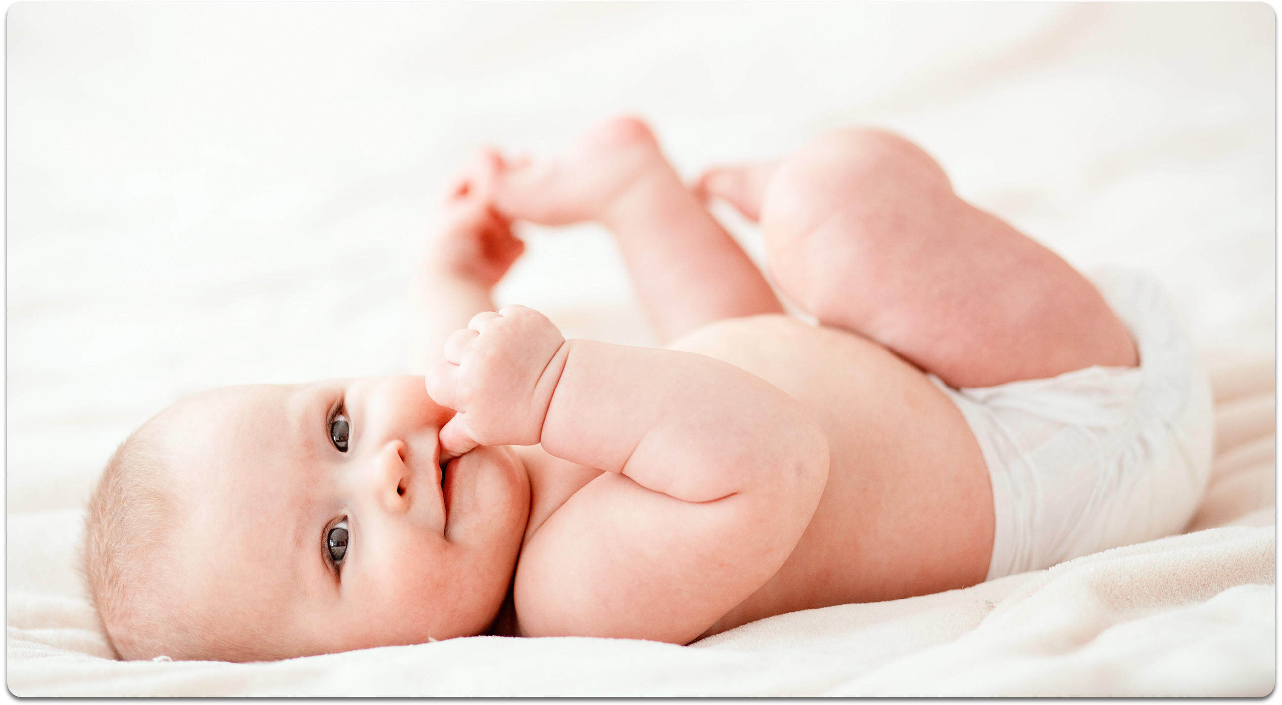 Baby in Windel liegt auf heller Decke und hält die Füße mit den Händen.