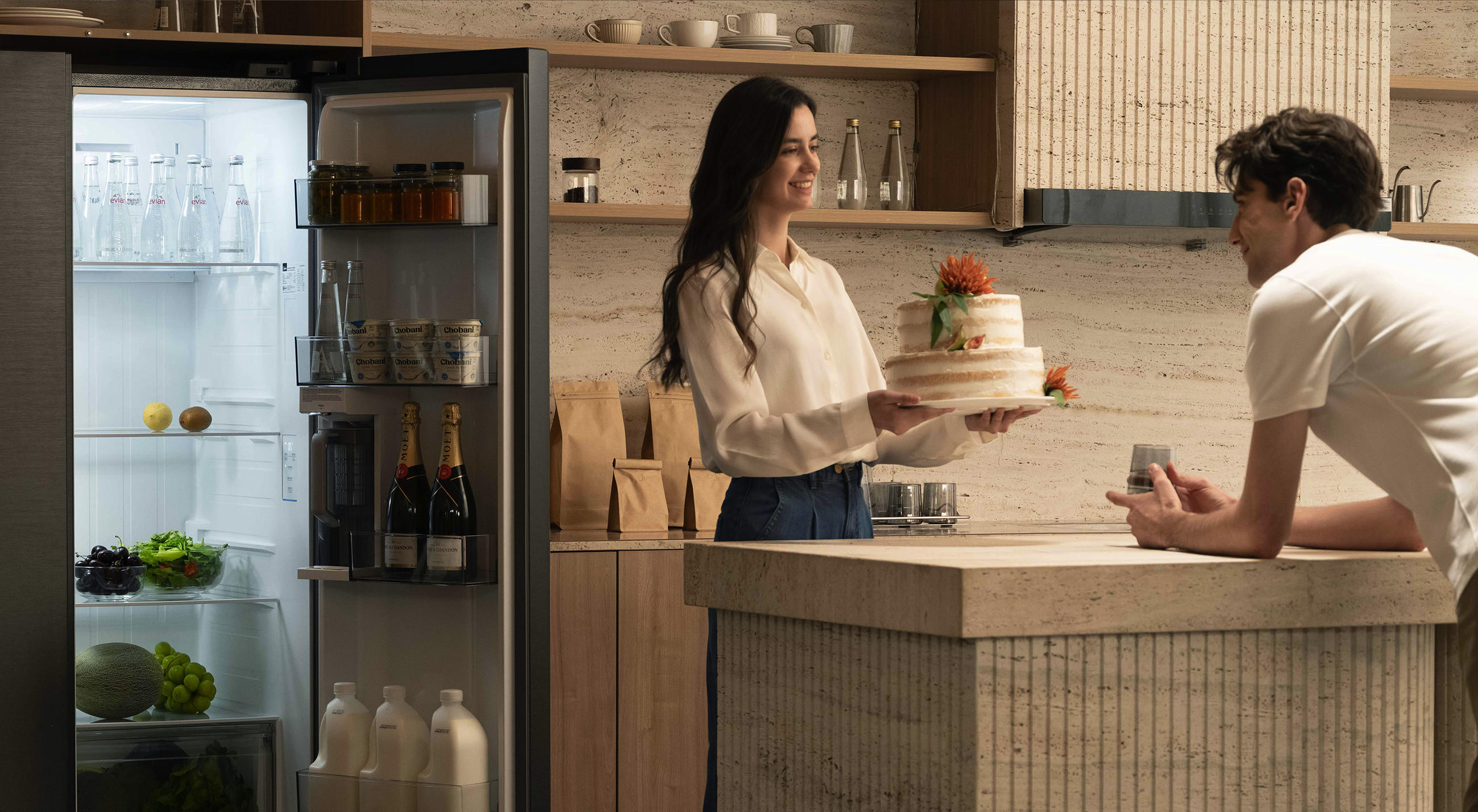 Offener Midea-Kühlschrank in einer modernen Küche, gefüllt mit Getränken und Lebensmitteln; daneben hält eine Frau eine Torte, während ein Mann an der Kücheninsel sitzt.