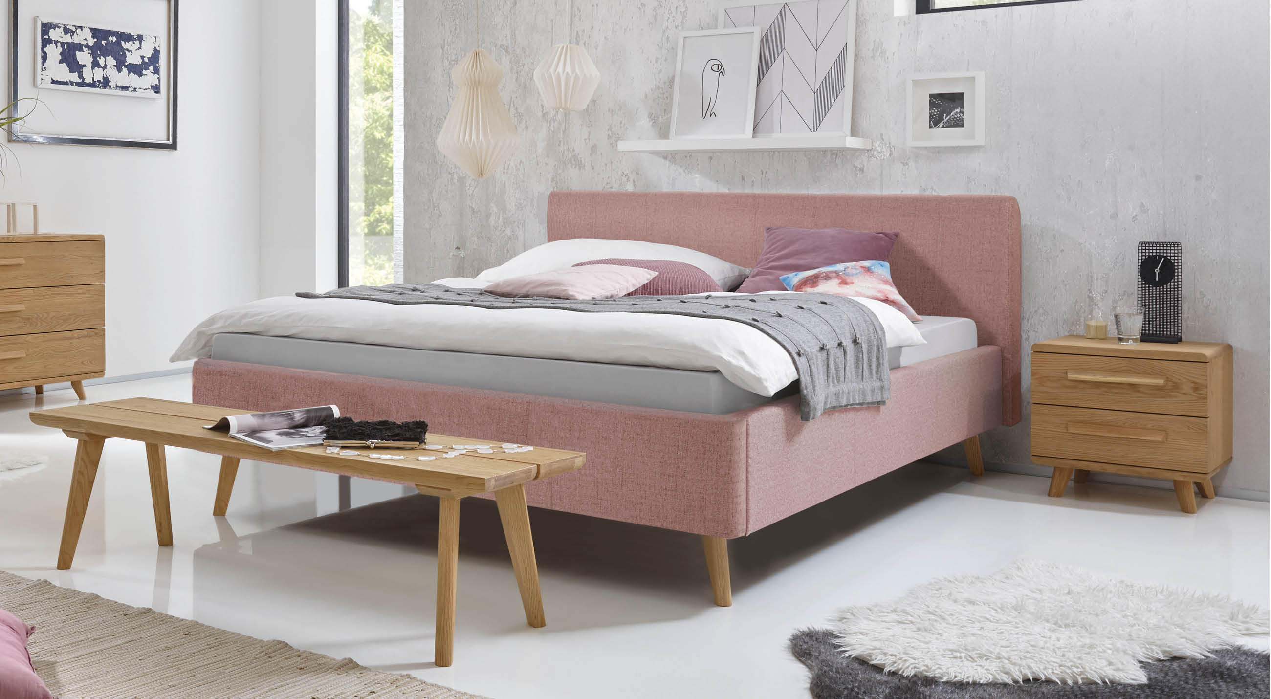 Hasena Polsterbett mit roséfarbenem Stoffbezug und geradlinigem Kopfteil, kombiniert mit Holzmöbeln und dekorativen Leuchten in einem hellen, modern eingerichteten Schlafzimmer.