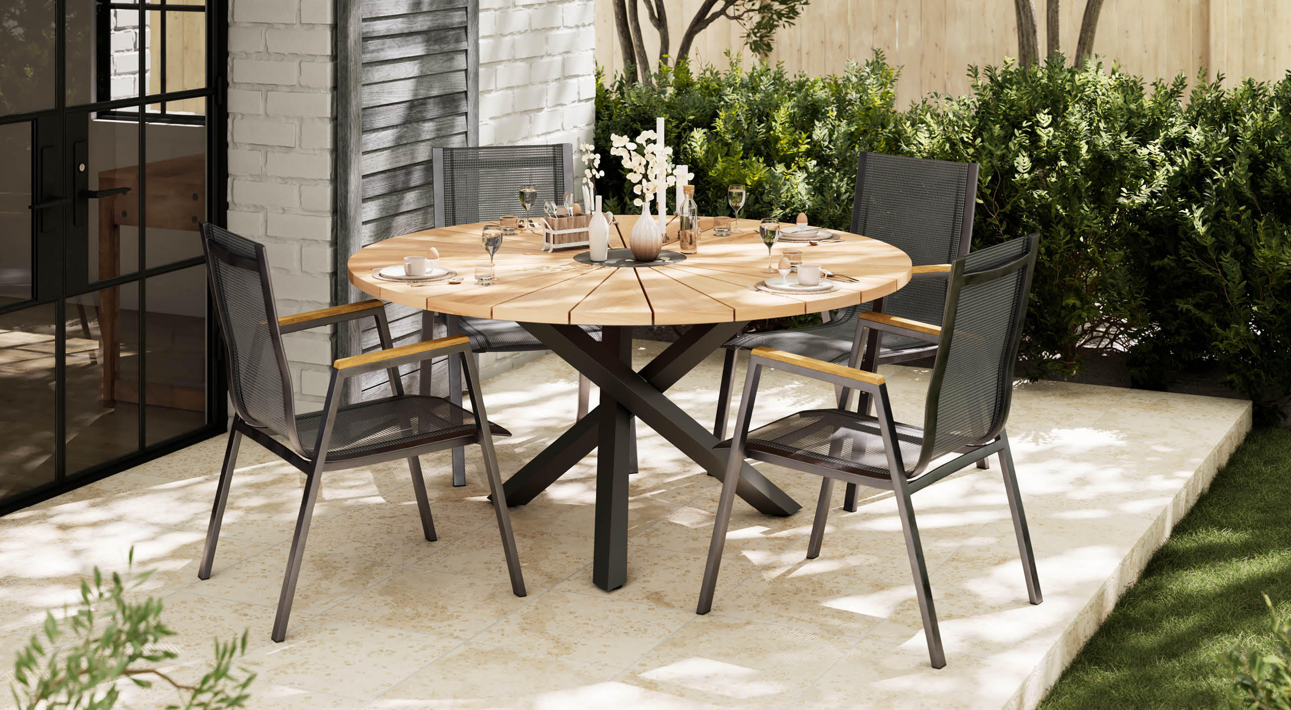 Runder Holzgartentisch mit vier grauen Stühlen auf einer Terrasse, gedeckt mit Geschirr und Vasen, Hecke im Hintergrund.