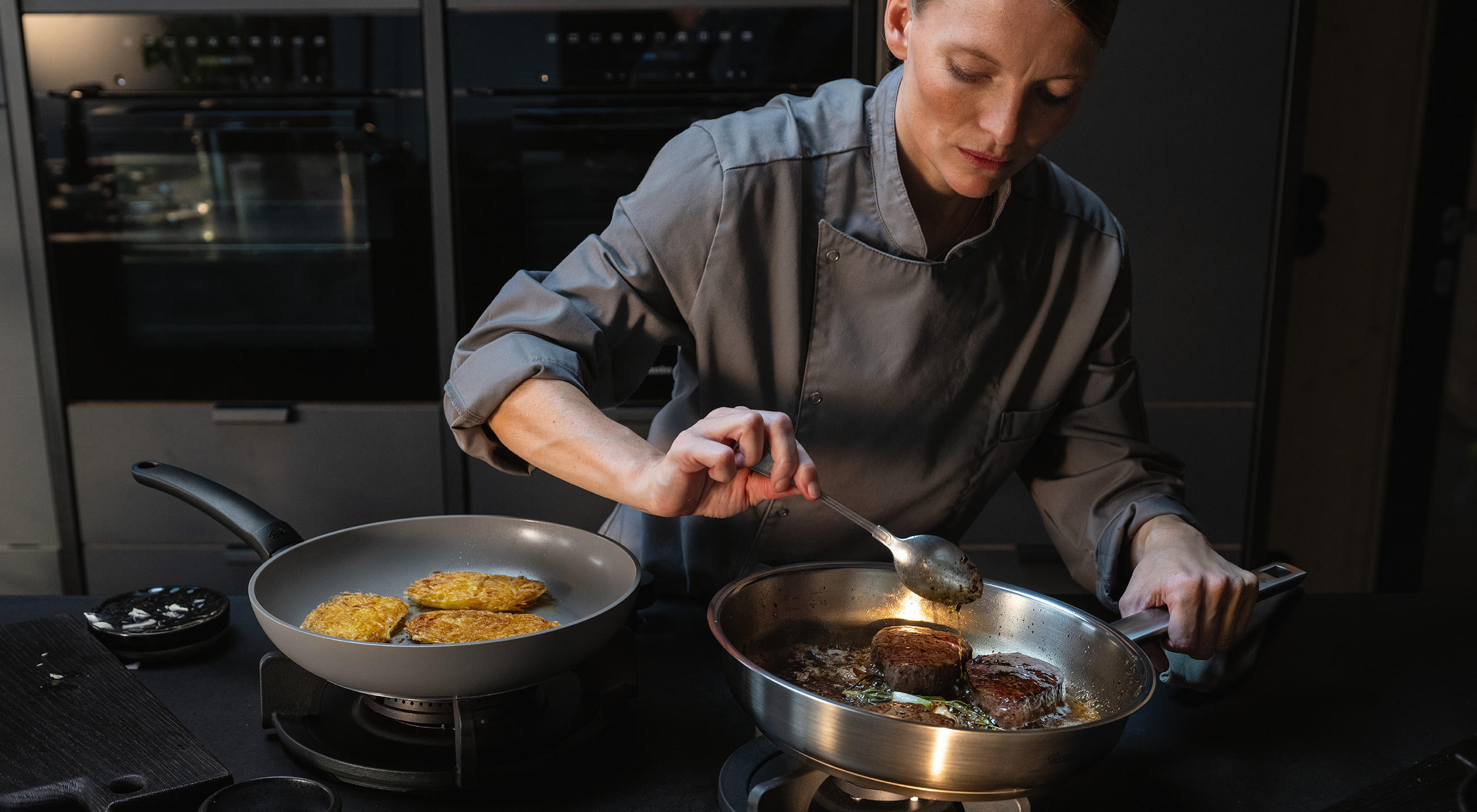 Köchin in grauer Kochjacke brät in einer FISSLER Edelstahlpfanne zwei Fleischstücke und übergießt sie mit Bratensaft; daneben bräunen Rösti in einer weiteren Pfanne auf dem Herd.