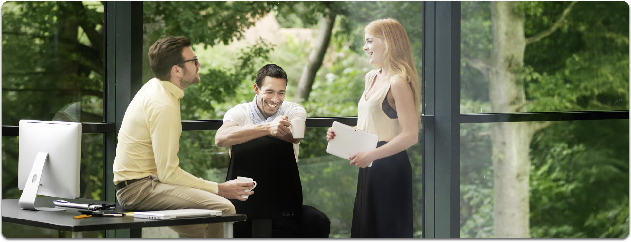 Drei Personen sitzen an einem Tisch mit einem Laptop und lachen gemeinsam; im Hintergrund ist grüner Wald durch die Glasfront zu erkennen.