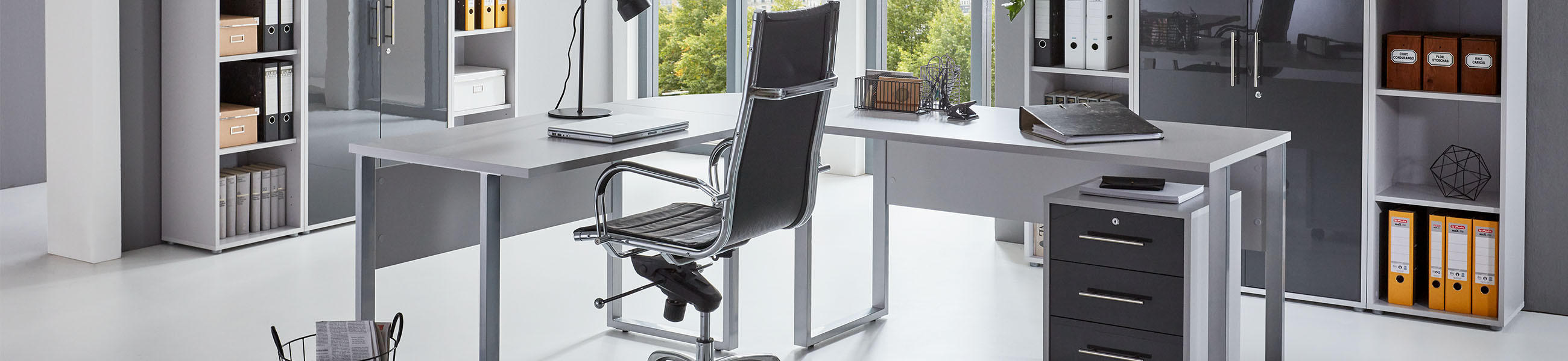Modernes Büro mit weißem Eckschreibtisch, schwarzem Bürostuhl, Rollcontainer und weißen anthrazit Aktenschränken vor großen Fenstern.
