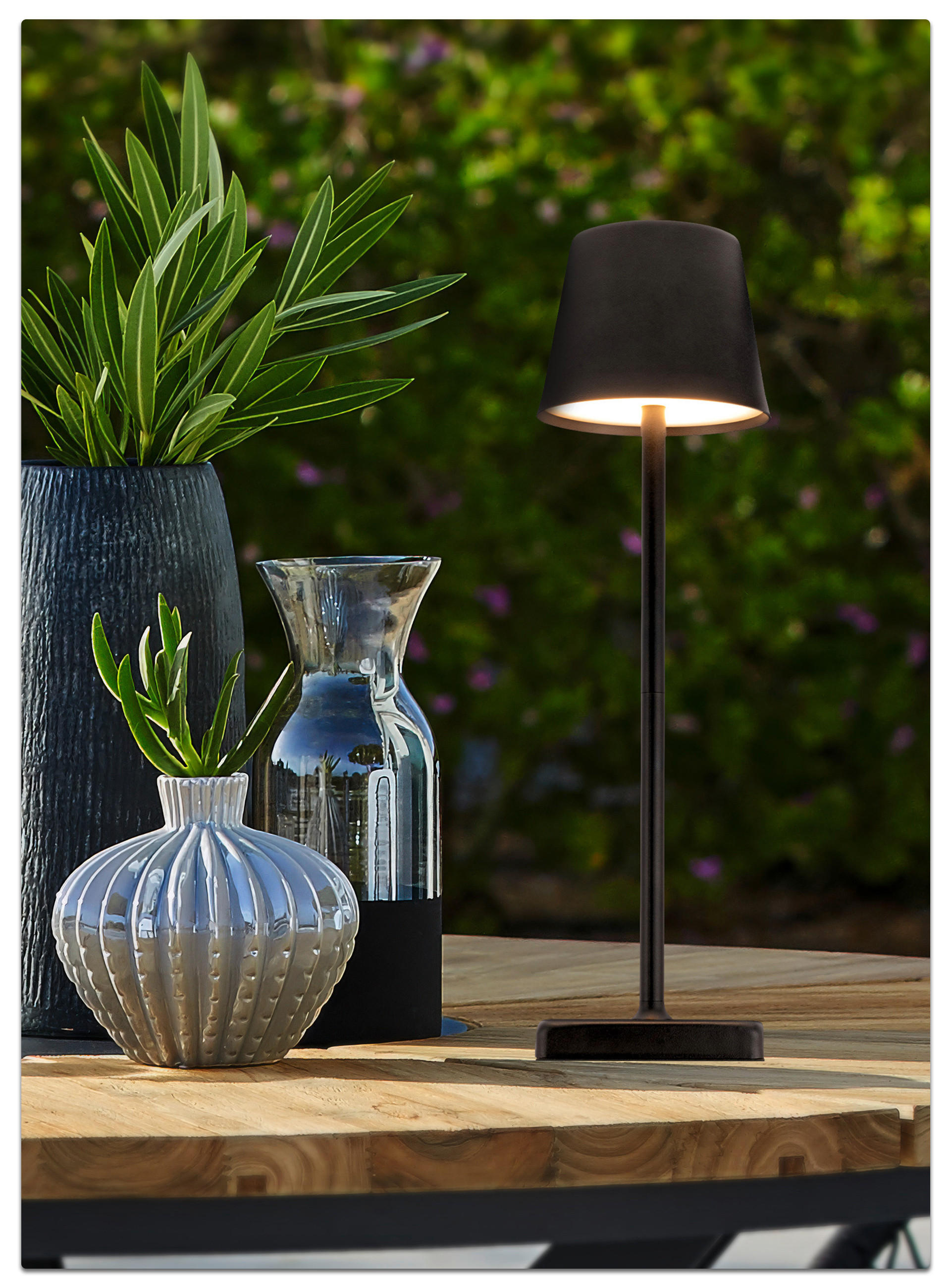 Holztisch mit beleuchteter Lampe, Vasen und grüner Dekoration vor blühendem Hintergrund.