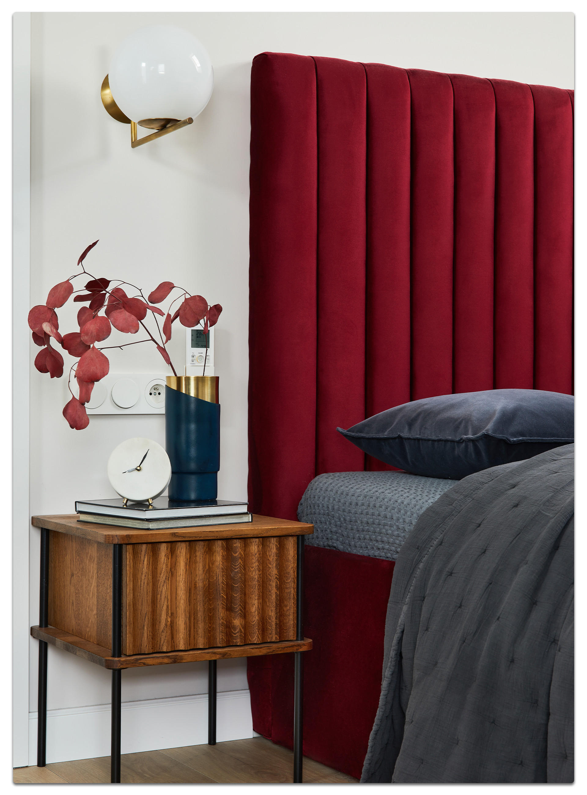 Schlafzimmer-Szene mit rotem Polsterkopfteil, blauer Vase mit roten Blättern auf Holz-Nachttisch, moderner Wandlampe und dunkler Bettwäsche – stilvolle Einrichtung mit warmen Farbakzenten.