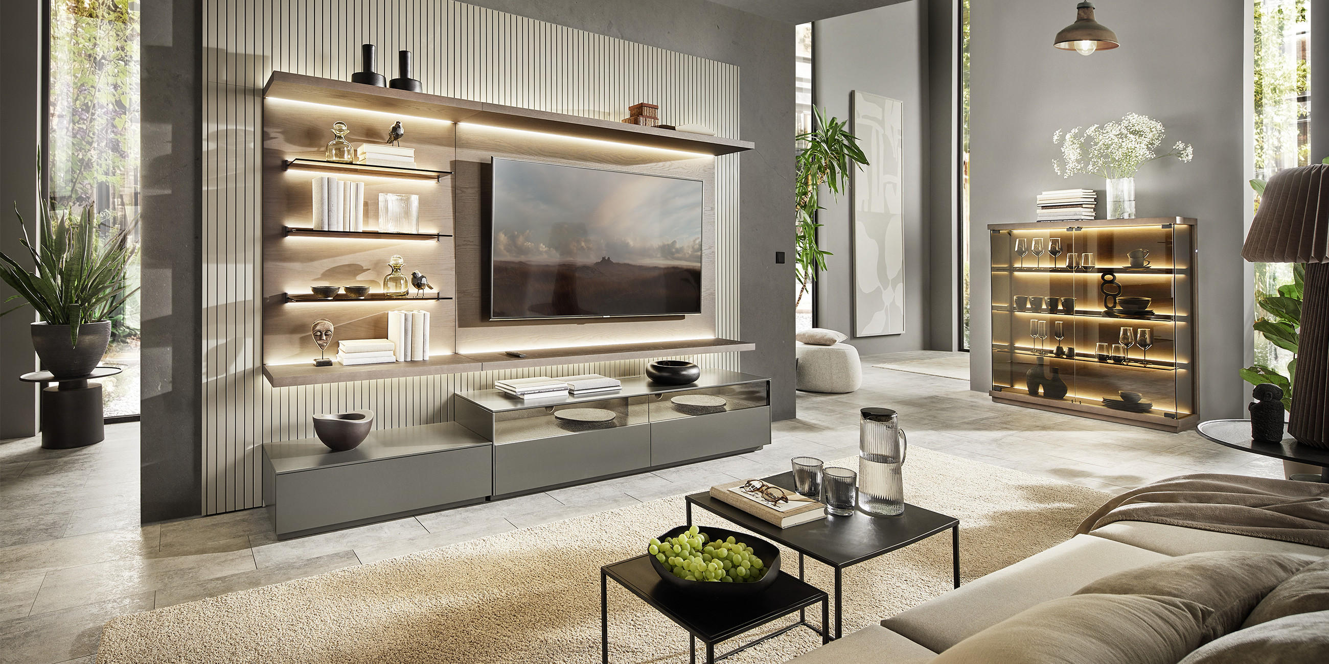 Modernes Wohnzimmer mit BELLUTI Sofa im Vordergrund und eleganter BELUTTI Wohnwand mit TV, beleuchteten Glasvitrinen und Regalen; dezente Deko in warmen Naturtönen.