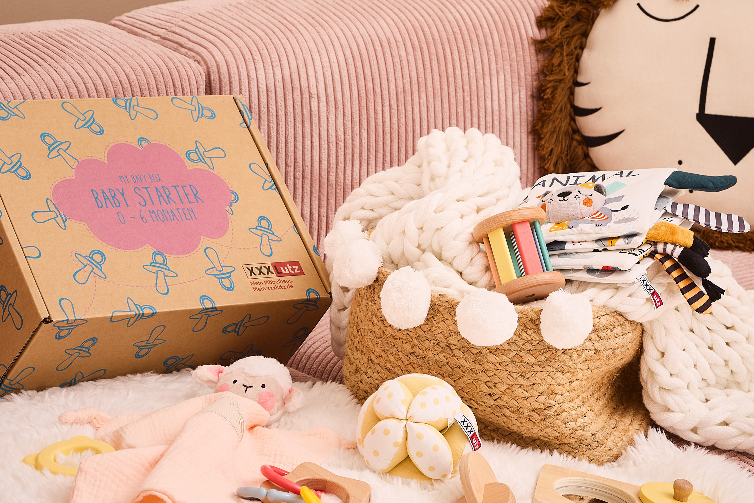 Baby-Starter-Box für 0–6 Monate von XXXLutz neben einem Korb mit weißer Strickdecke, Holzrassel und Stoffbüchern mit Tiermotiven. Im Hintergrund rosa Kissen und ein Dekokissen mit Tiergesicht