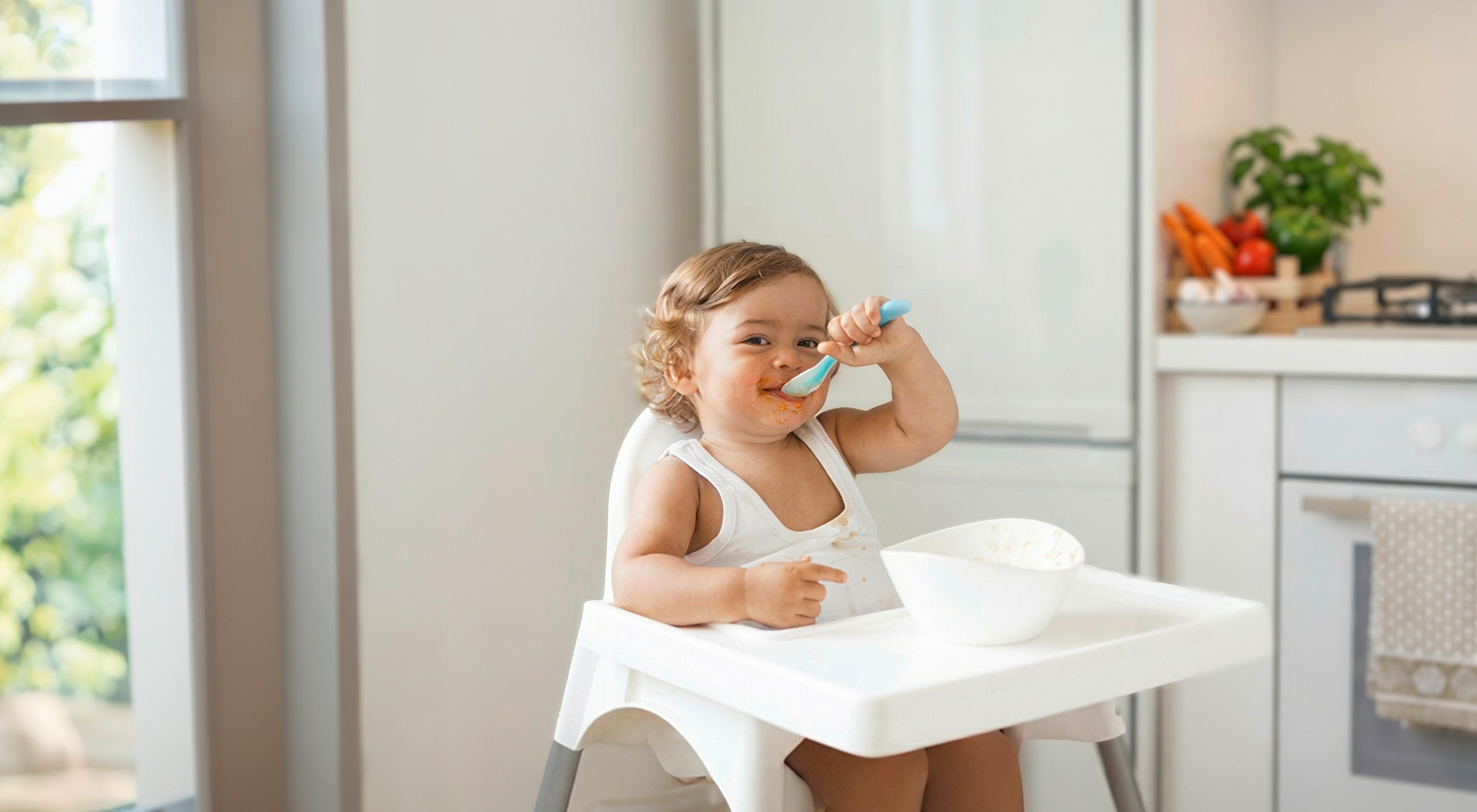 Ein Kleinkind sitzt in einem Hochstuhl in einer hellen Küche und isst aus einer Schüssel, während es einen Löffel in der Hand hält. Im Hintergrund stehen Gemüse und Küchenutensilien auf der Arbeitsfläche.