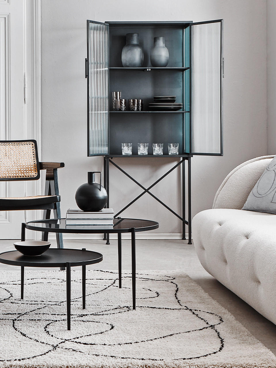 Modern eingerichteter Wohnbereich mit schwarzem Metall-Couchtisch, Sessel mit geflochtener Rückenlehne und Glasgeschirr in offenem Schrank, mit dem Hinweis „SHOP THE LOOK“