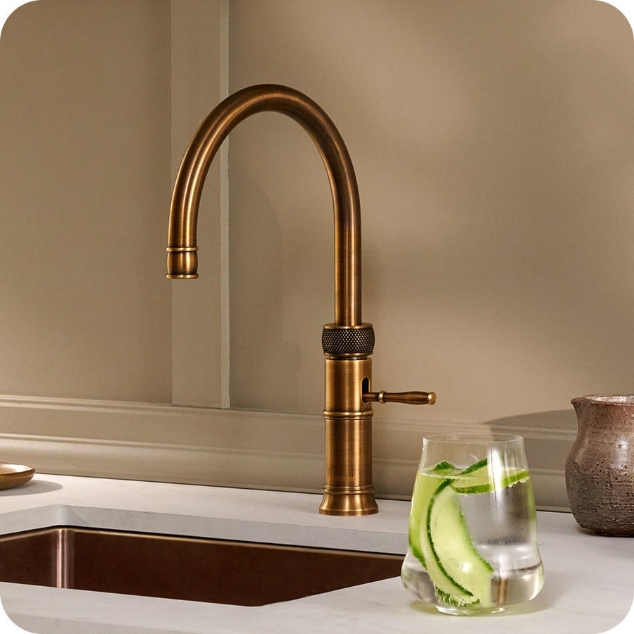 QUOOKER Classic Fusion Round Armatur in Messingoptik mit gebogenem Auslauf und Seitenhebel an einer hellen Spüle; daneben ein Glas Wasser mit Gurkenscheiben auf der Arbeitsplatte.