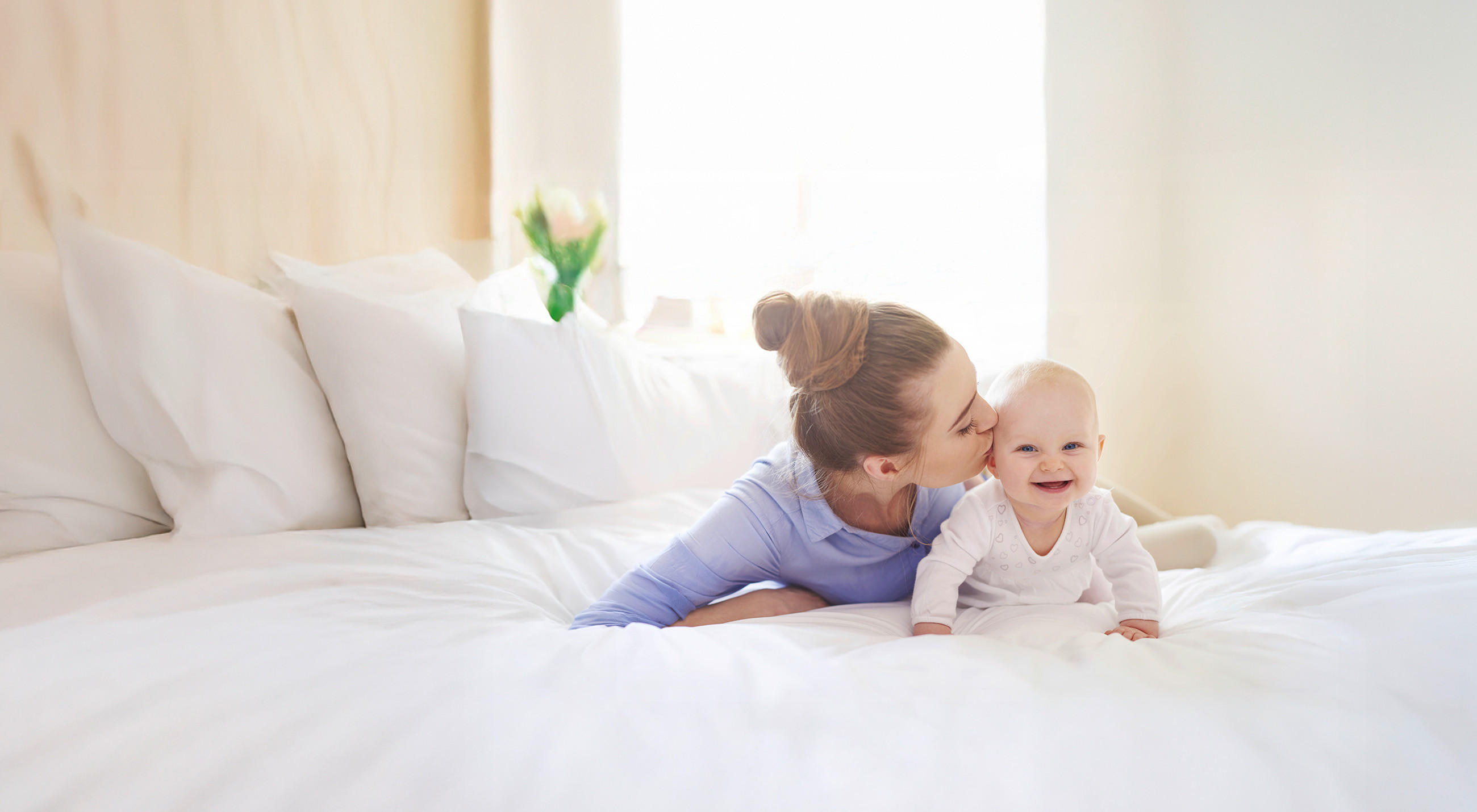 Eine Person liegt entspannt auf einem großen weißen Bett und stützt sich auf den Ellbogen, während ein Baby in weißer Kleidung vor ihr sitzt. Der Raum ist hell und freundlich, mit weichen Kissen und natürlichem Licht, das durch ein Fenster einfällt.