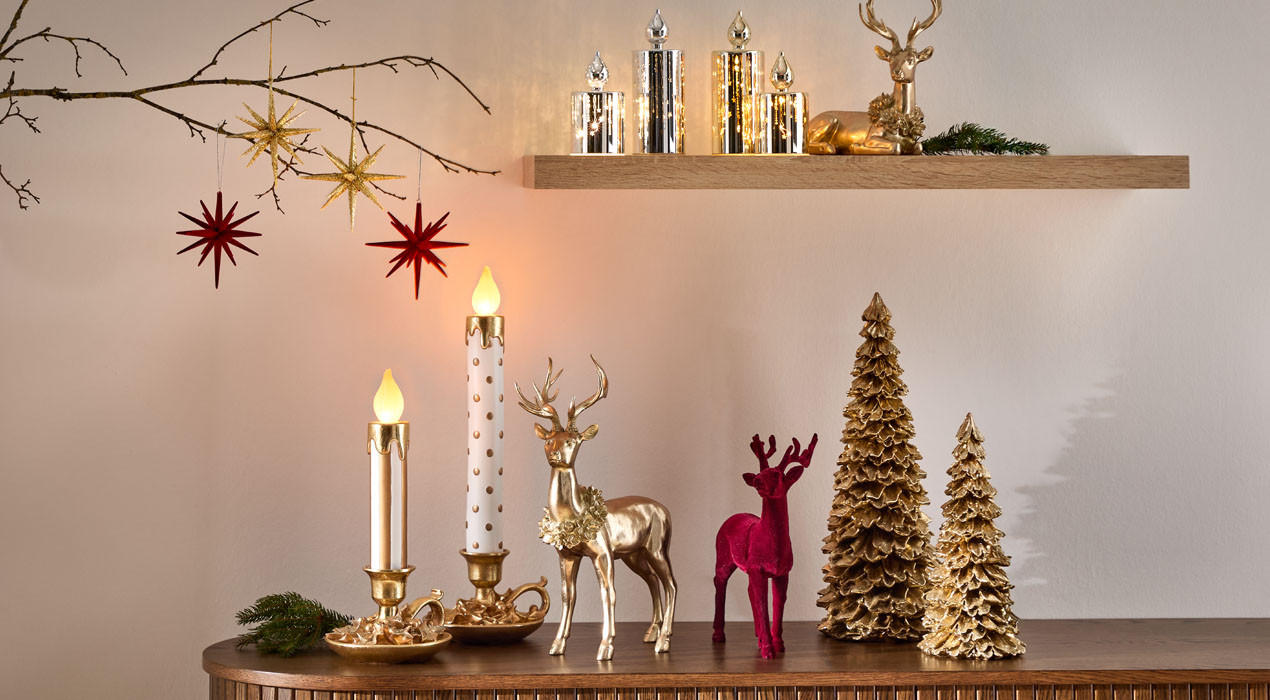Festliche Szene mit Kerzen, goldenen und roten Rentierfiguren sowie goldenen Weihnachtsbaum-Ornamenten auf einem Holztisch.