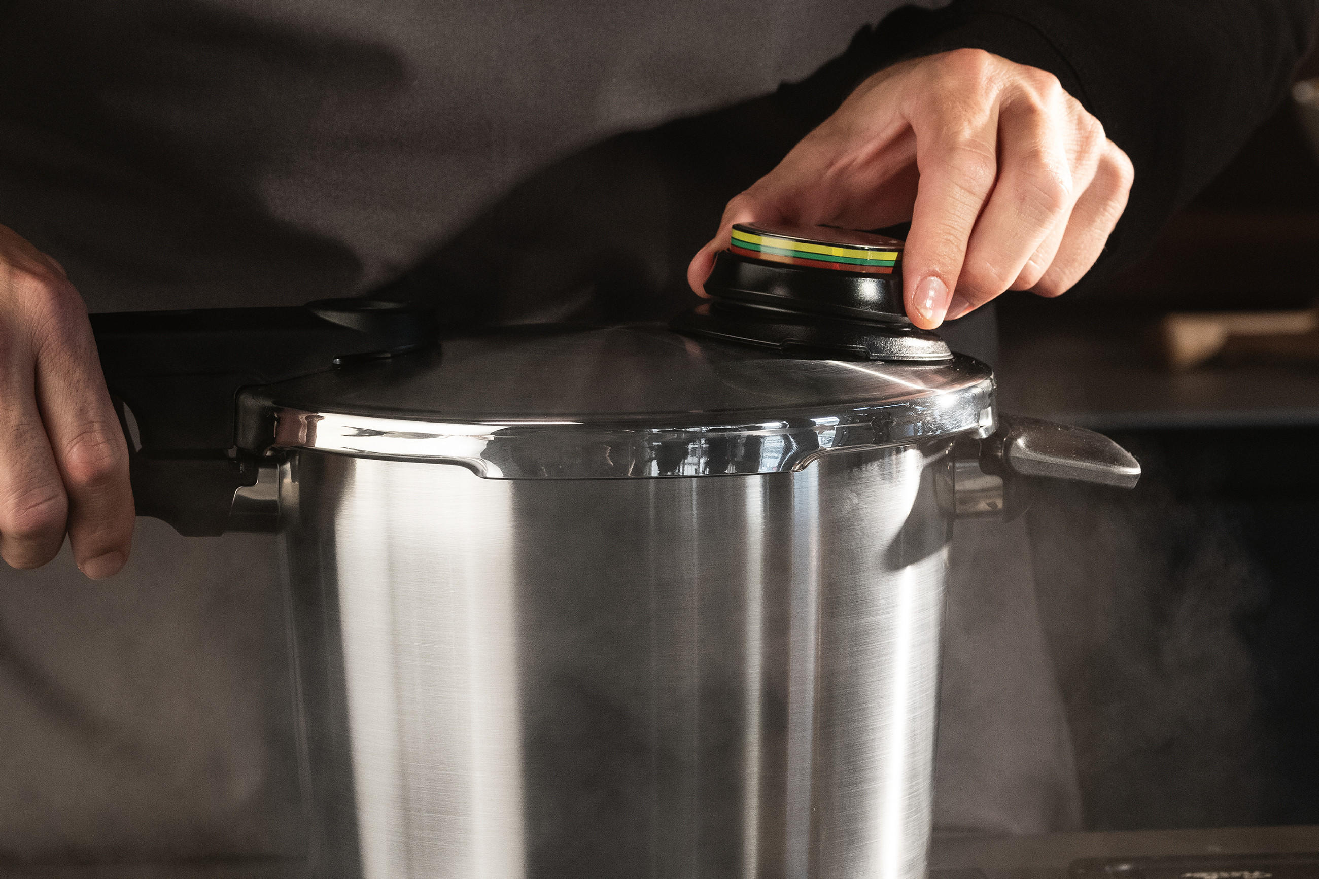 Nahaufnahme eines dampfenden Fissler Vitaquick® Premium Schnellkochtopfs aus Edelstahl; Hände bedienen den Deckelgriff und die farbige Druckanzeige oben am Ventil.