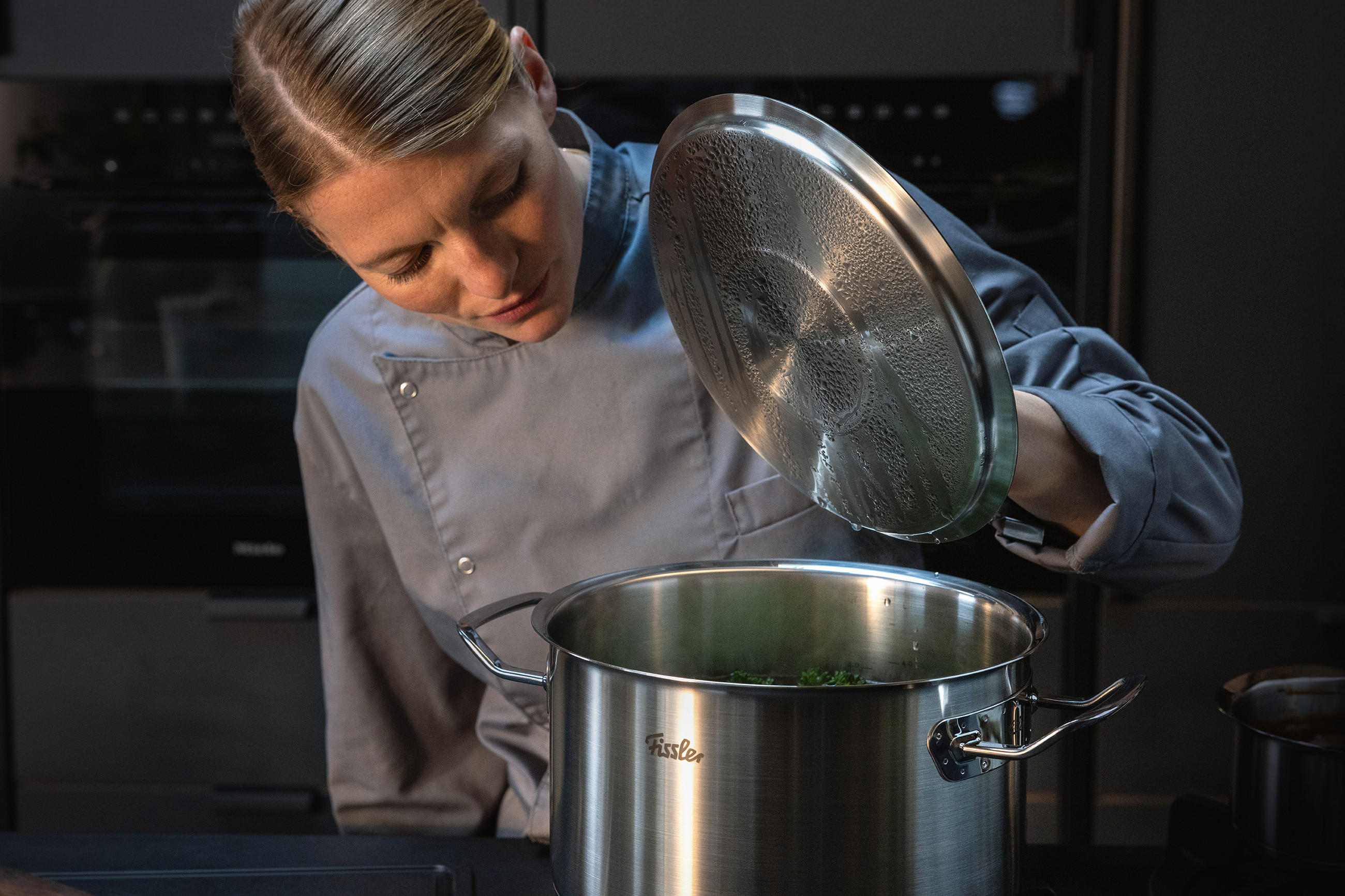 Köchin in grauer Kochjacke hebt den beschlagenen Deckel eines großen FISSLER Edelstahl‑Topfs an und blickt in den Topf; das Markenlogo ist auf der Topfwand sichtbar, im Hintergrund eine dunkle Profiküche.