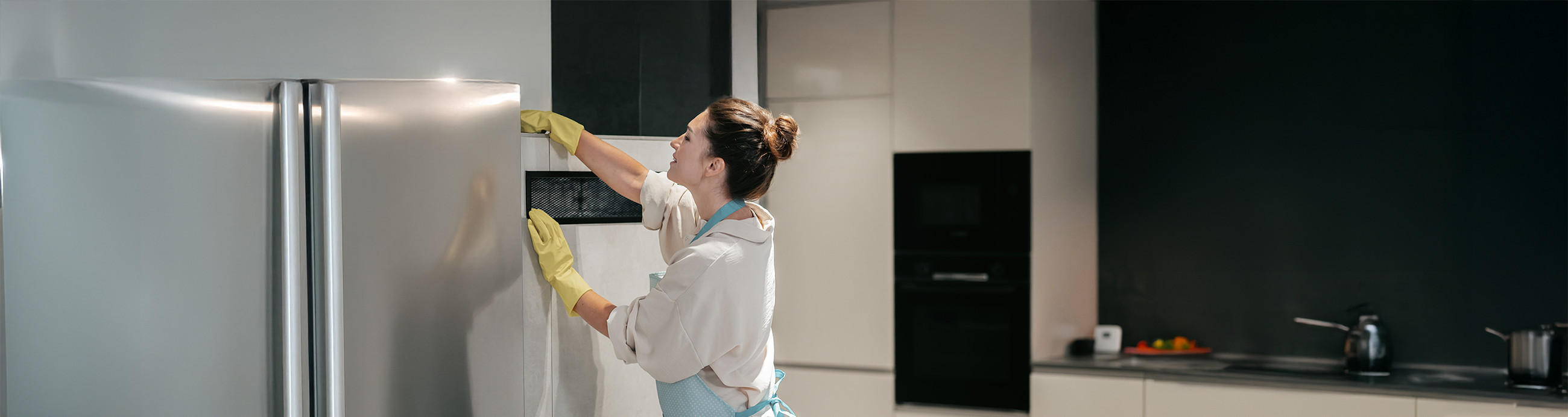 Eine Frau in Handschuhen und Schürze steht in moderner Küche und reinigt einen Kühlschrank von außen.