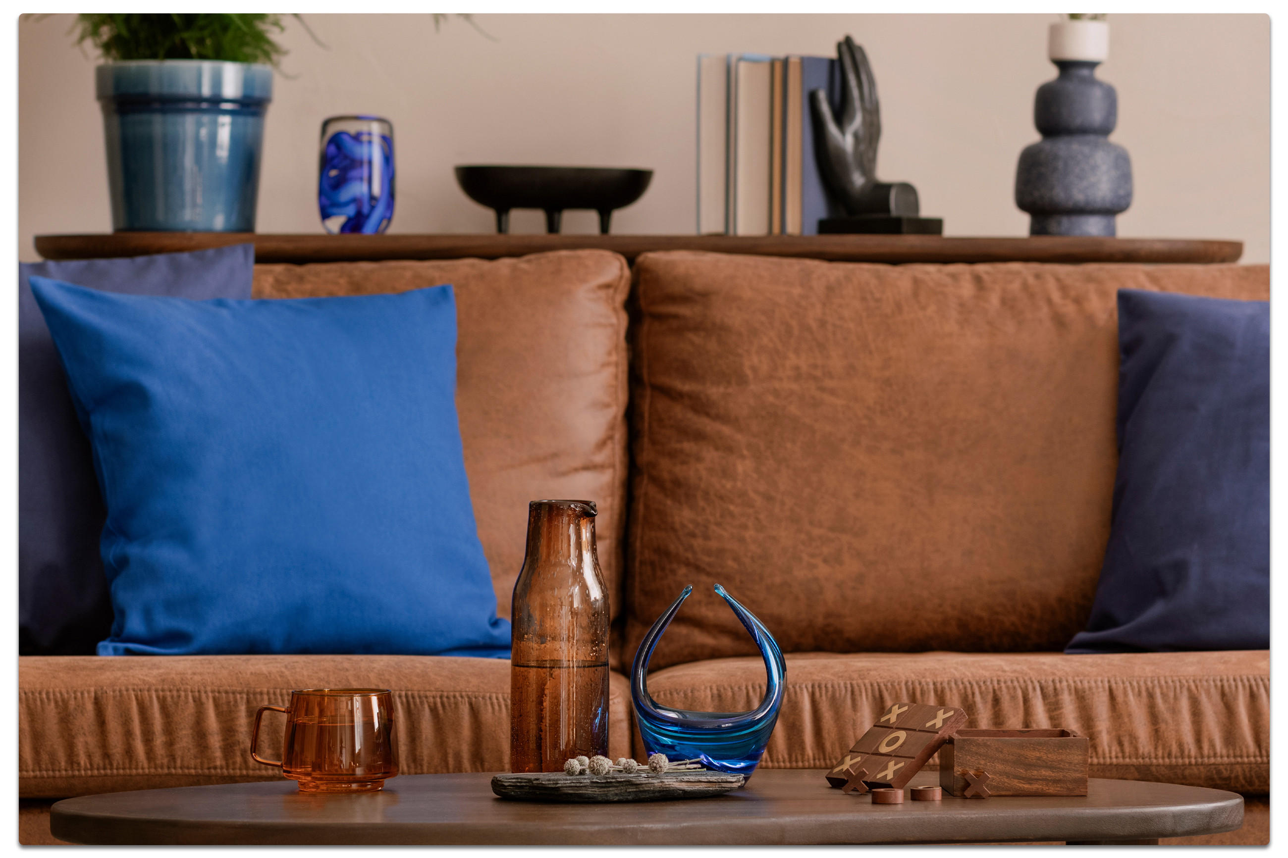 Modernes Wohnzimmer mit braunem Ledersofa, blauen Kissen, Holzcouchtisch mit Dekoobjekten und abstraktem Gemälde in Blau – stilvolle Einrichtung mit wohnlichem Flair.