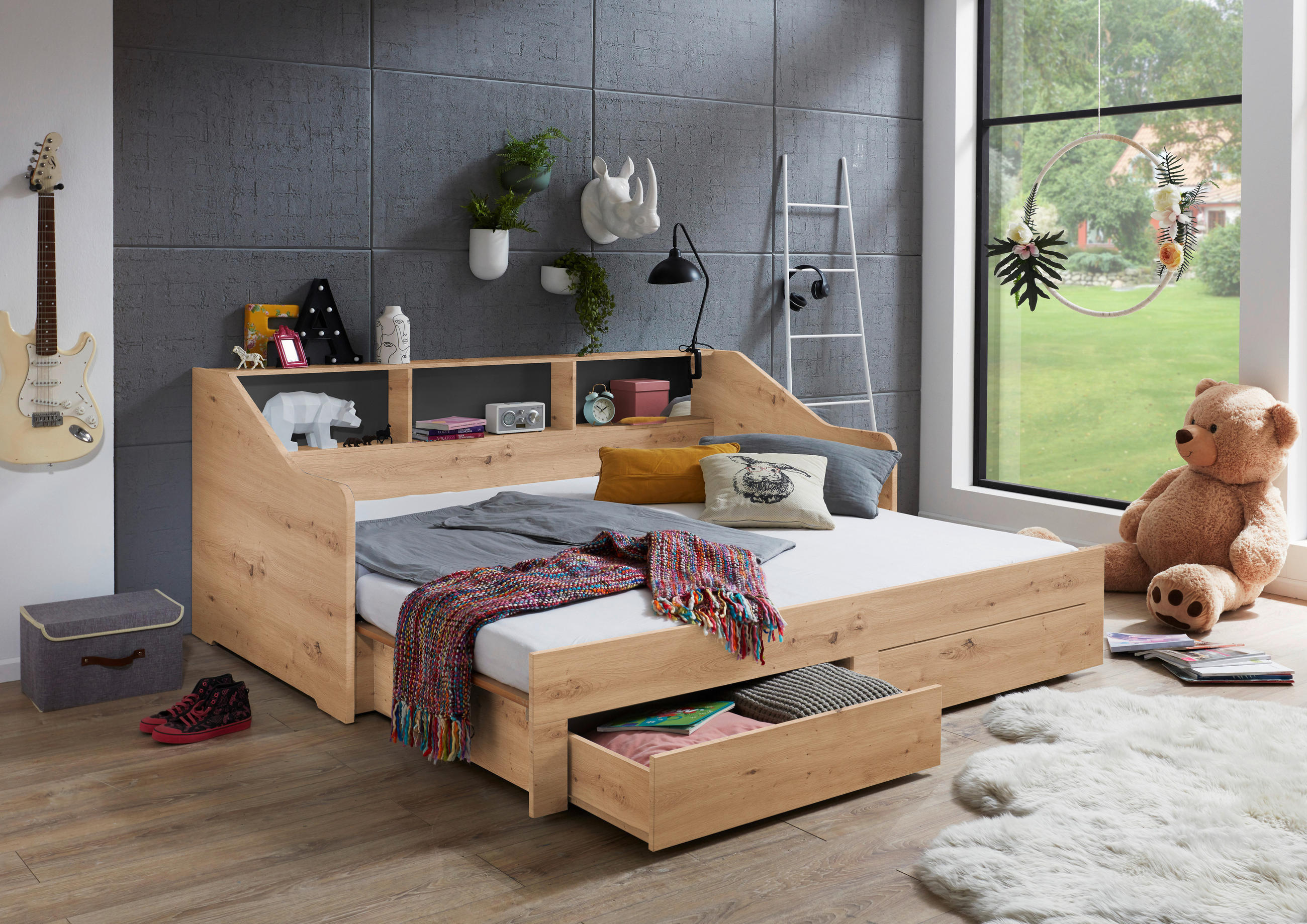 Funkcionalna enojna postelja iz svetlega lesa z integriranimi policami v vzglavju in dvema spodnjima predaloma za shranjevanje. Postelja je raztegljiva in se lahko spremeni v dvojno ležišče, idealno za mladinske ali otroške sobe.