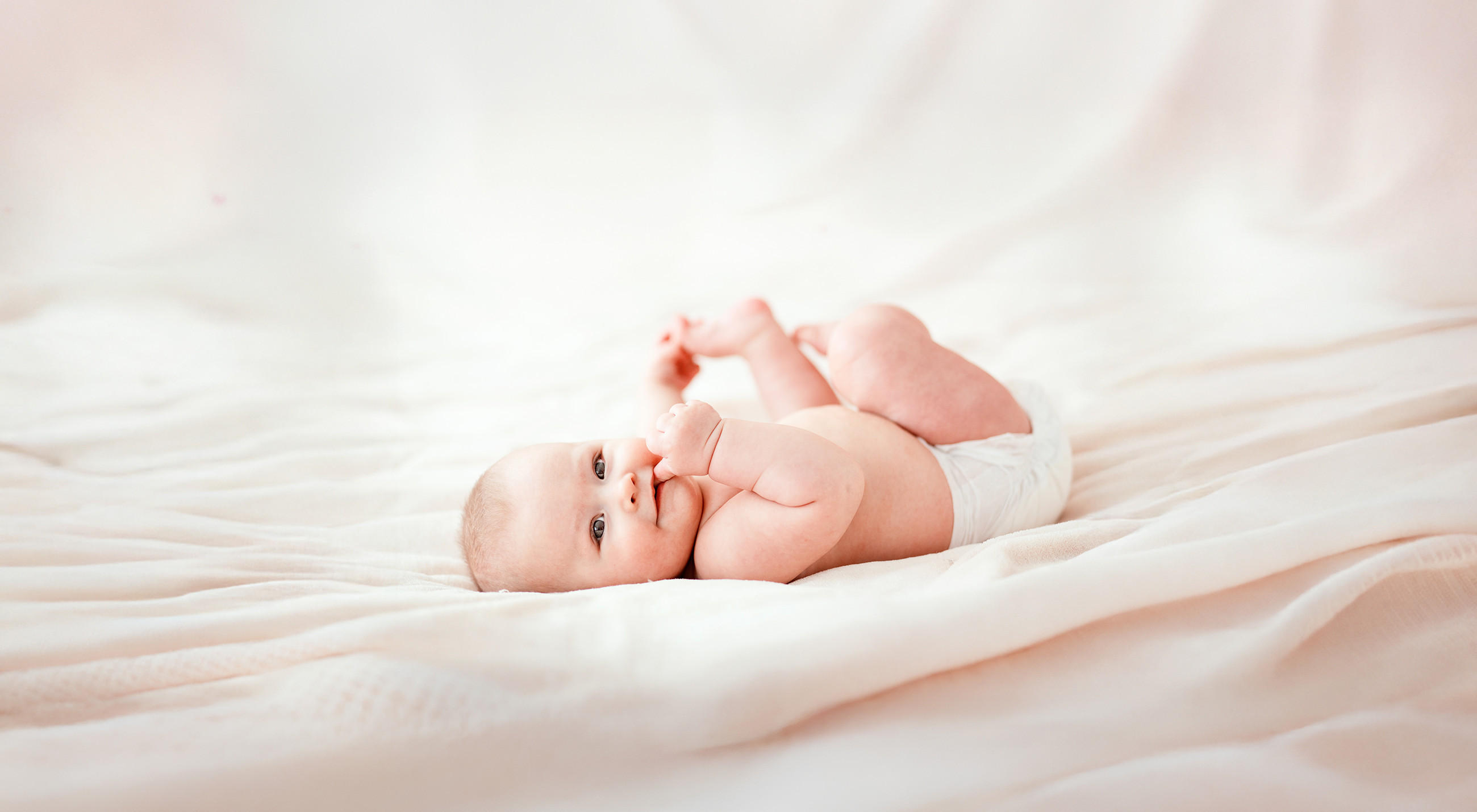 Ein Baby liegt auf dem Rücken auf weichen, hellen Decken und hebt spielerisch Arme und Beine in die Luft. The setting wirkt warm und sanft, mit weichem Licht und einem beruhigenden, cremefarbenen Hintergrund. 