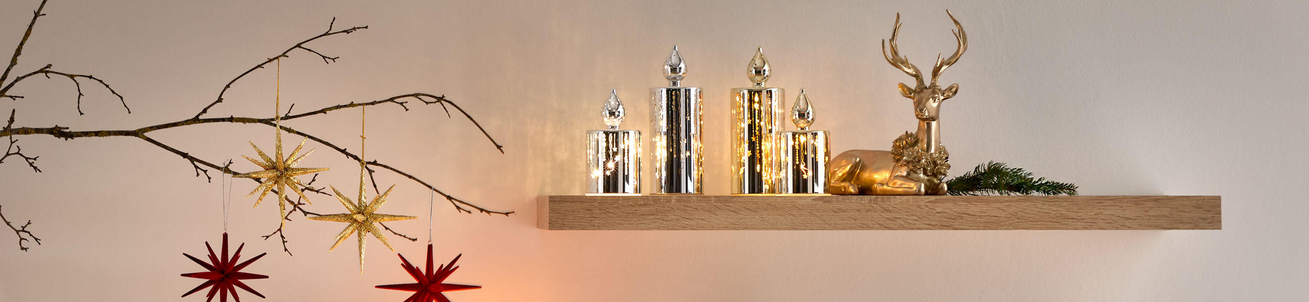 Dekoratives Arrangement mit vier Kerzenhaltern, roten und goldenen Sternanhängern sowie einem liegenden goldenen Hirsch auf einem Holzregal.