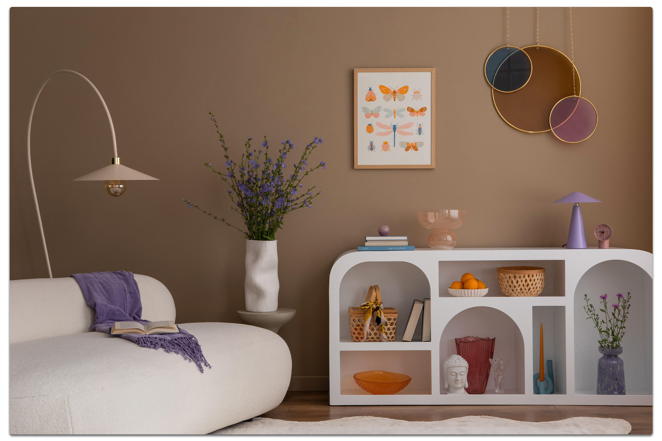 tilvolles Wohnzimmer mit weißem Sofa und lila Decke, weißer Vase mit lila Blumen, dekorativem Regal und Wandkunst mit Schmetterlingen und Spiegeln – gemütliches Ambiente mit warmen Brauntönen.