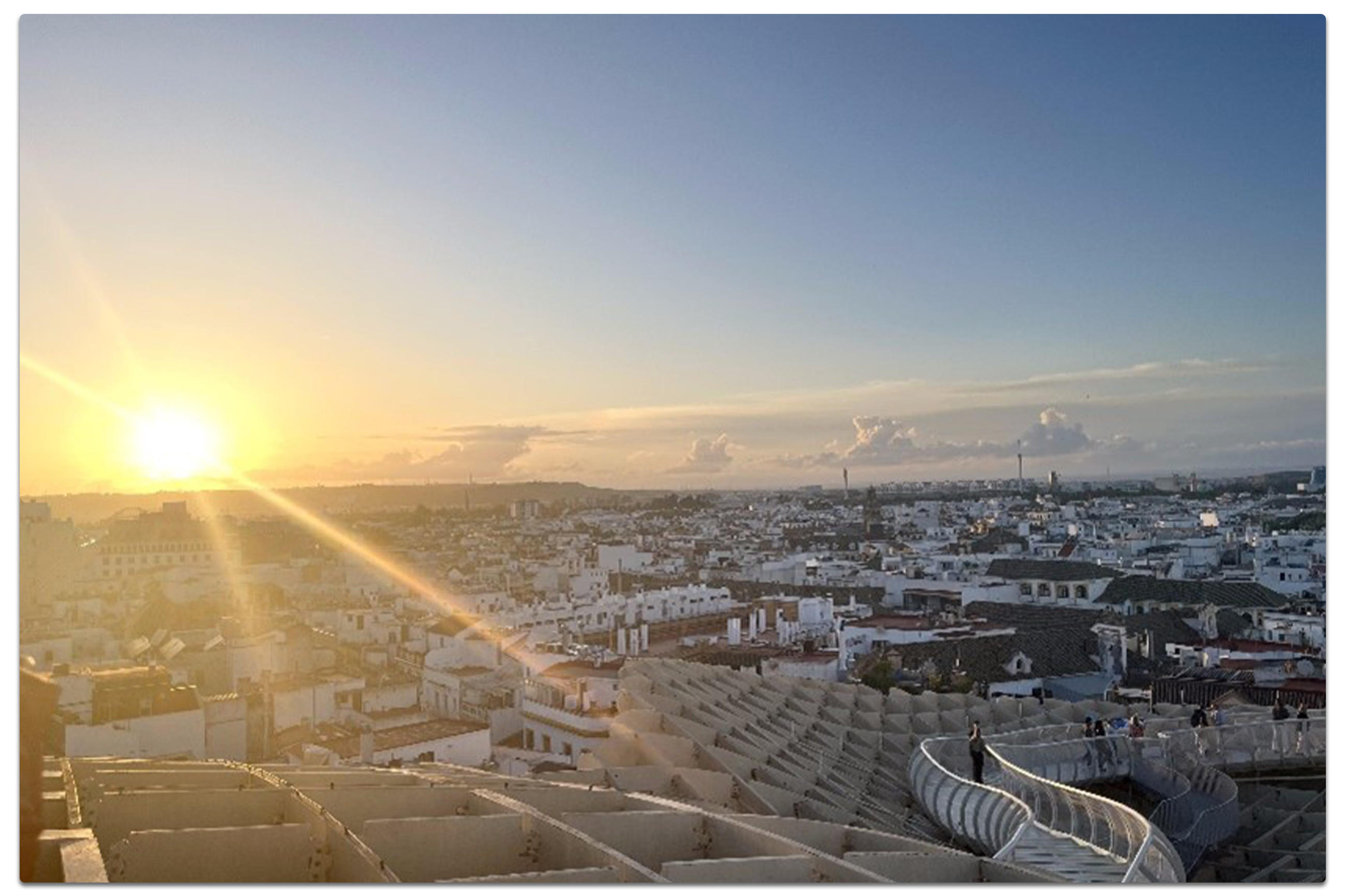 Panoramablick auf eine spanische Stadt bei Sonnenuntergang mit geschwungenem Architektur-Element im Vordergrund und goldener Lichtstimmung über den Dächern.