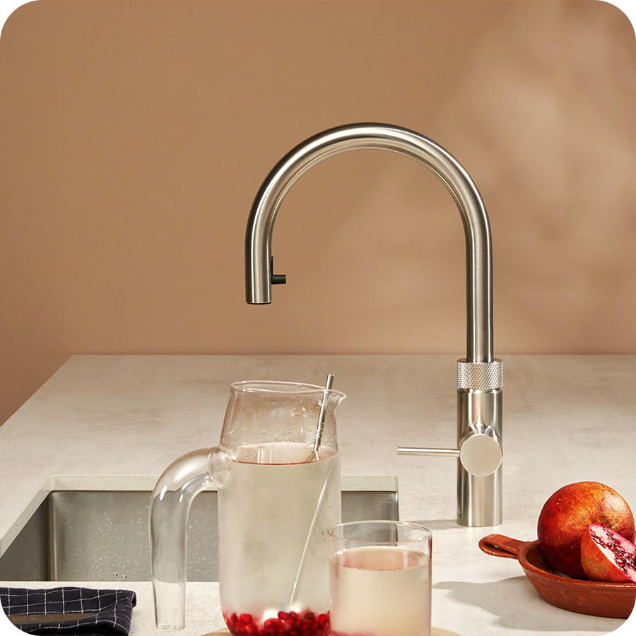 QUOOKER Fusion Round; Edelstahl-Küchenarmatur mit hohem Rundbogen über einer Spüle; auf der Arbeitsfläche stehen eine Glaskaraffe und ein Glas mit Getränk sowie Granatapfelstücke, im Hintergrund ein warm getönter Wandbereich.