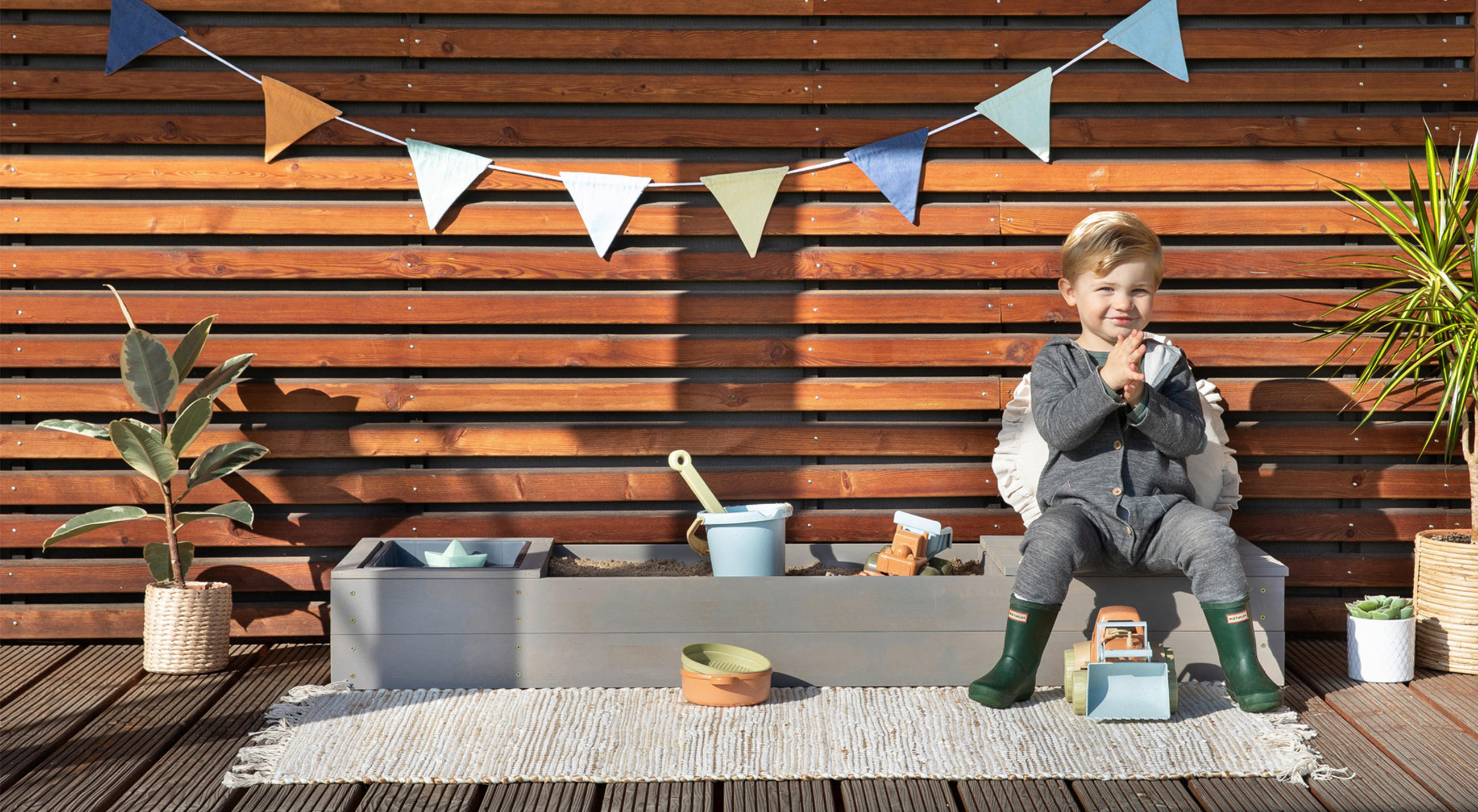 Kind mit Gummistiefeln sitzt auf einer Holzbank neben Pflanze und Spielzeug vor einer Holzlattenwand mit dem Schriftzug; XXXLUTZ BABYWELT RATGEBER