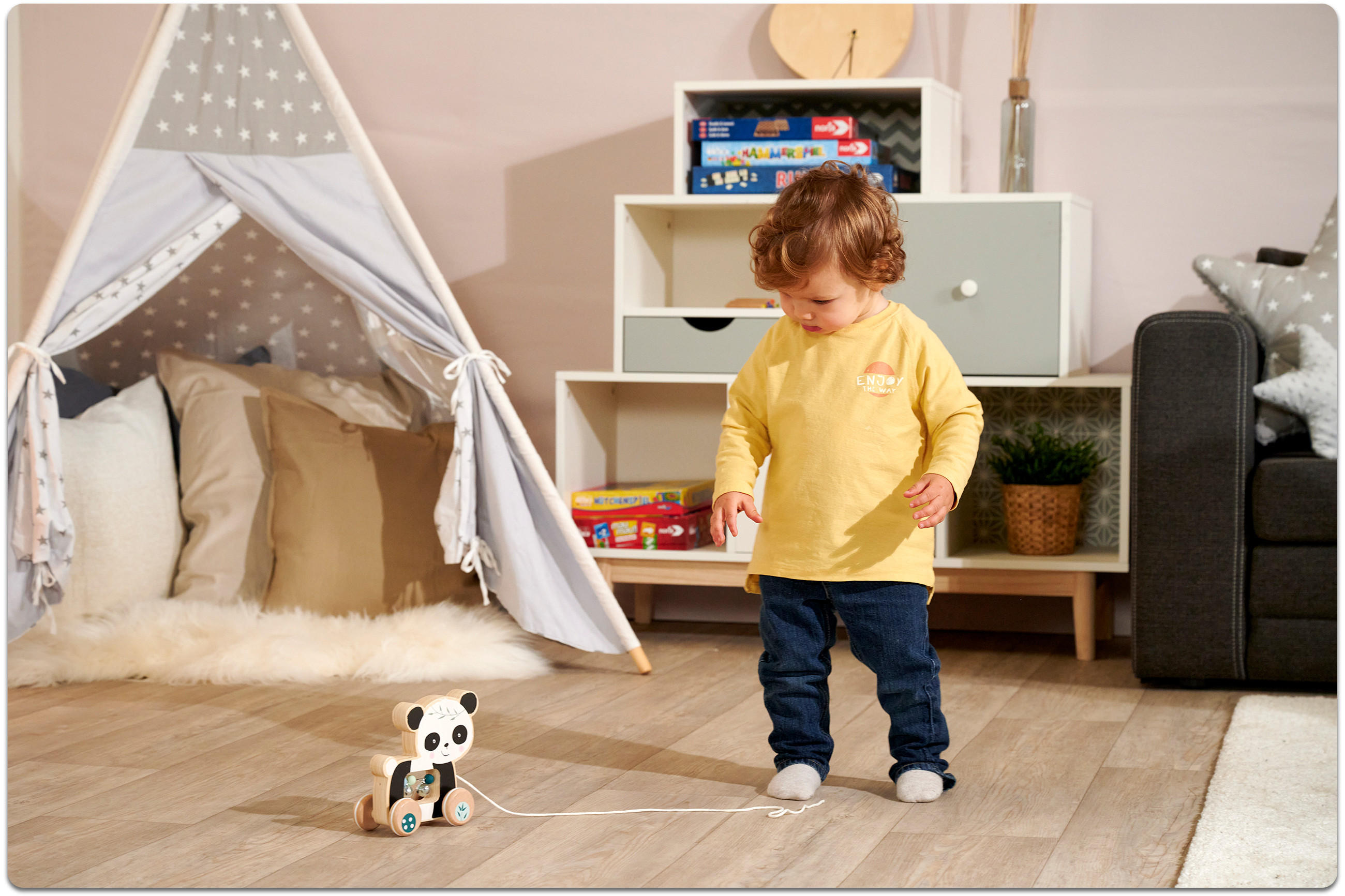 Kleinkind mit gelbem Langarmshirt und Jeans steht auf hellem Holzboden vor einem Spielzelt und einem weißen Regal mit Spielzeug und Pflanze.