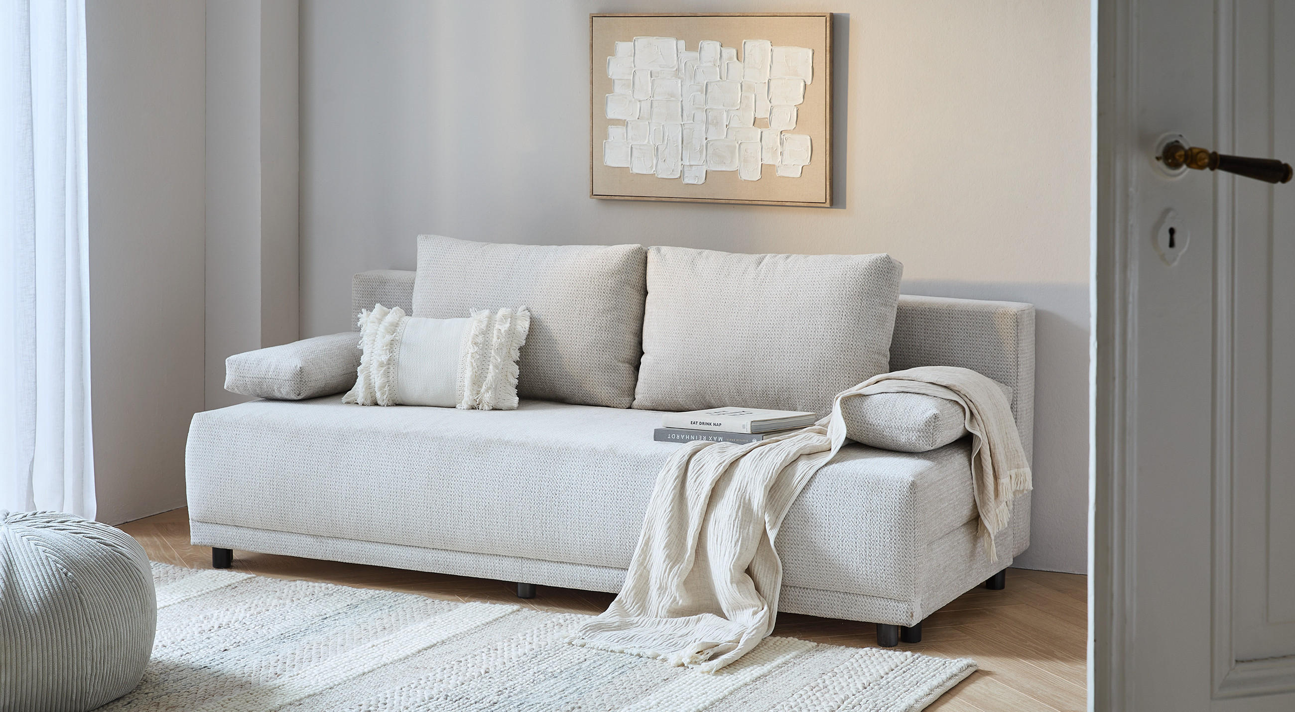 Hellgraues Sofa mit zwei Kissen und einer Decke in einem hellen Wohnzimmer mit Teppich und Wandbild.