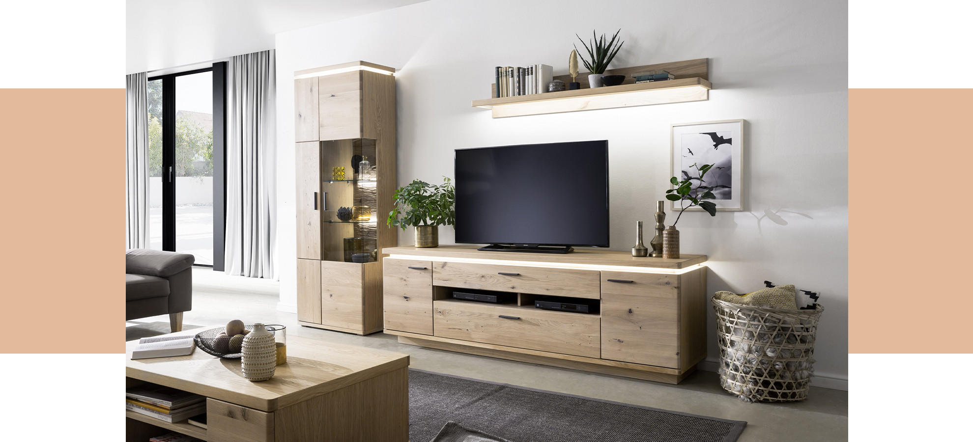Wohnzimmer mit hellem Holzsideboard, großem Flachbildfernseher, Vitrine, Couchtisch und Korb neben weißer Wand mit Regal und Bild