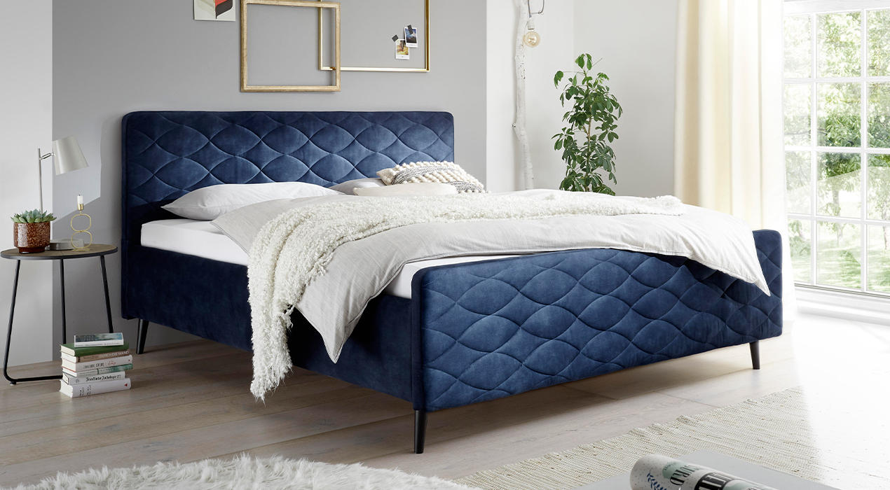 Blaues Polsterbett mit Oval-Steppung auf Kopf- und Fußteil. Weißer Topper, Bettdecke und Kissen.