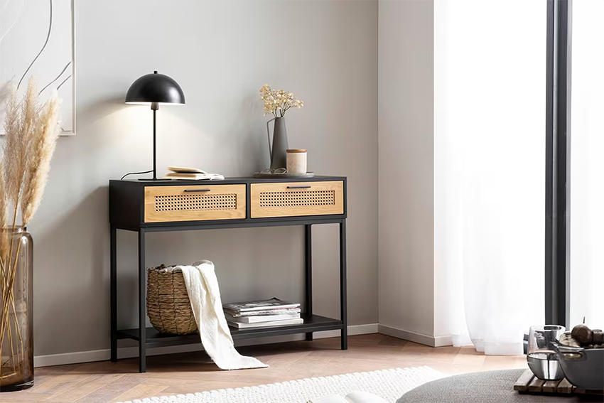 Modern konzolasztal fekete fém vázzal és rattan hatású fiókelőkkel, letisztult enteriőrben dekorlámpával és kiegészítőkkel.