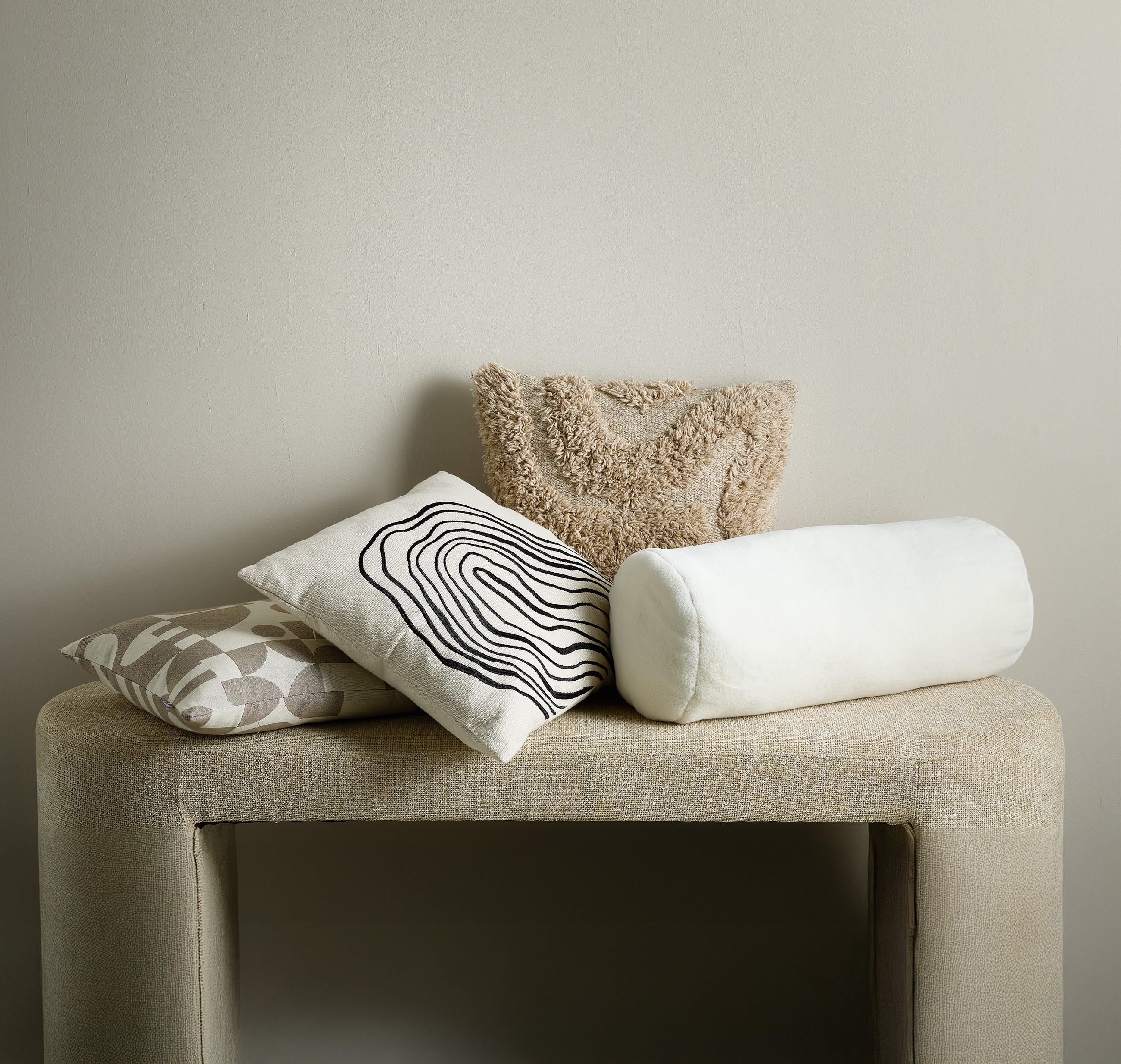 Vier dekorative Kissen in verschiedenen Formen und Texturen liegen auf einer hellen, gepolsterten Bank vor einer neutralen Wand.