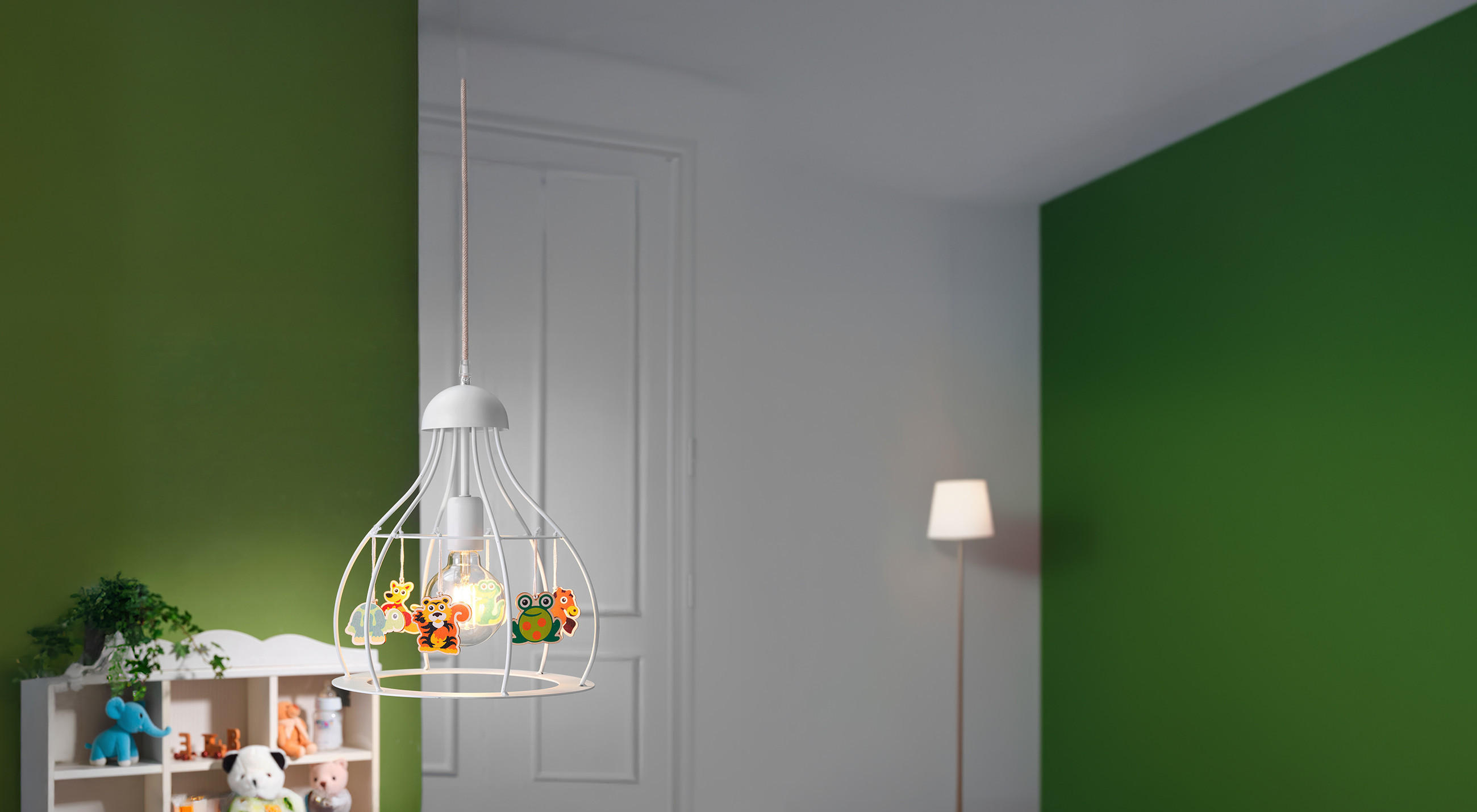 Eine dekorative Kinderzimmer‑Hängeleuchte mit weißem Metallgestell hängt im Raum; an ihr sind bunte Tierfiguren befestigt. Im Hintergrund stehen ein Regal mit Stofftieren, eine weiße Tür und eine schlanke Stehlampe vor einer grün‑weißen Wand.