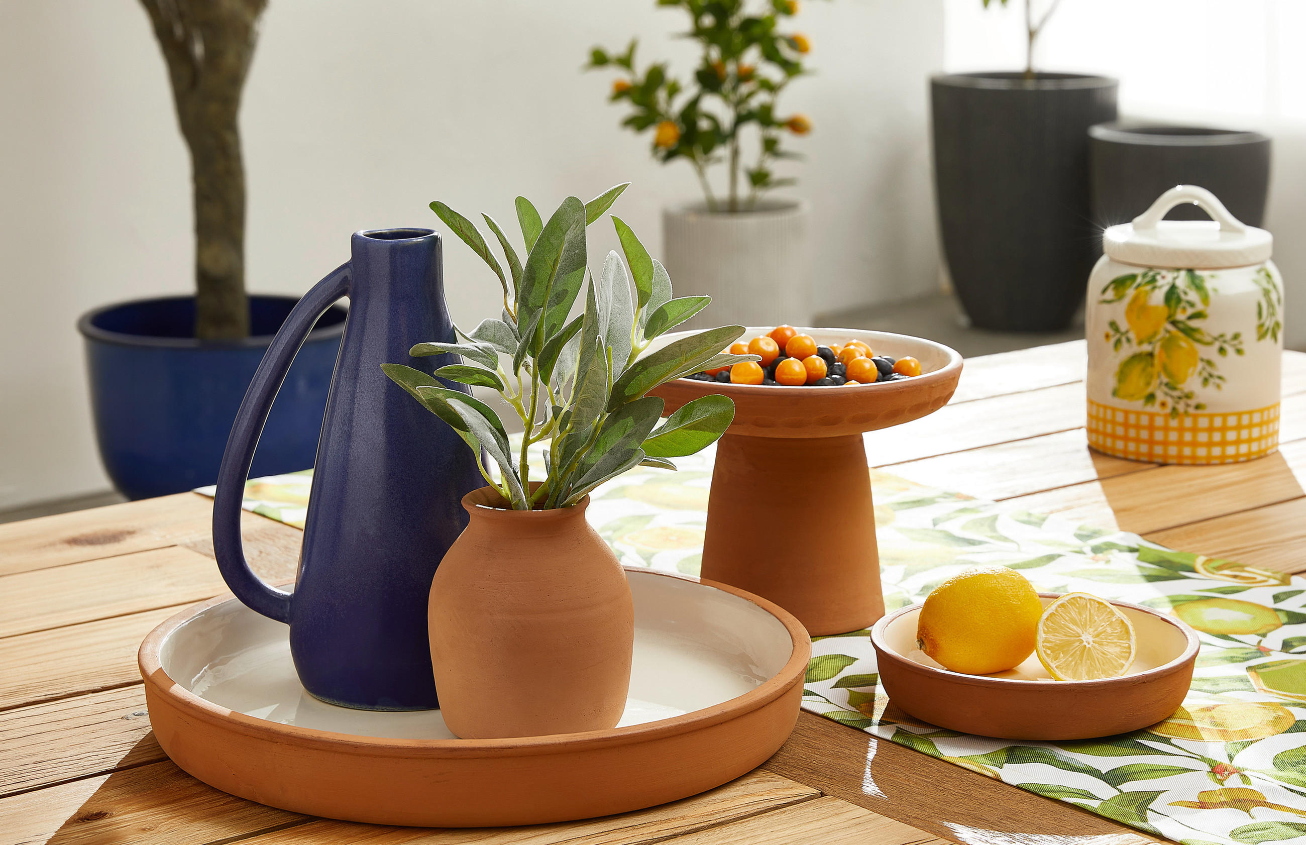 Tisch mit braunen Keramikgefäßen, darunter eine Vase mit grünen Blättern, eine blaue Kanne, eine Schale mit kleinen Orangenfrüchten und eine kleine Schale mit einer Zitrone.