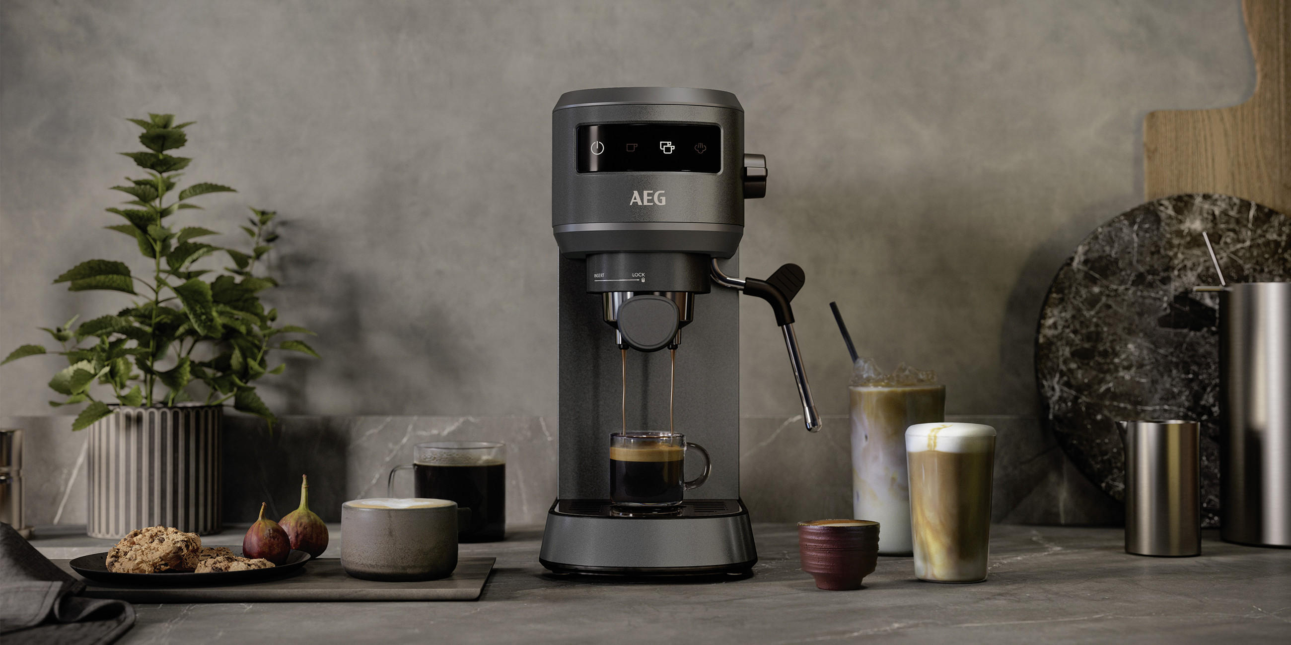 Kompakte AEG Espressomaschine auf dunkler Arbeitsplatte brüht Kaffee in eine Tasse; daneben stehen Latte-Gläser, Milchgetränke, Kekse, Feigen und eine Topfpflanze vor grauer Wand.