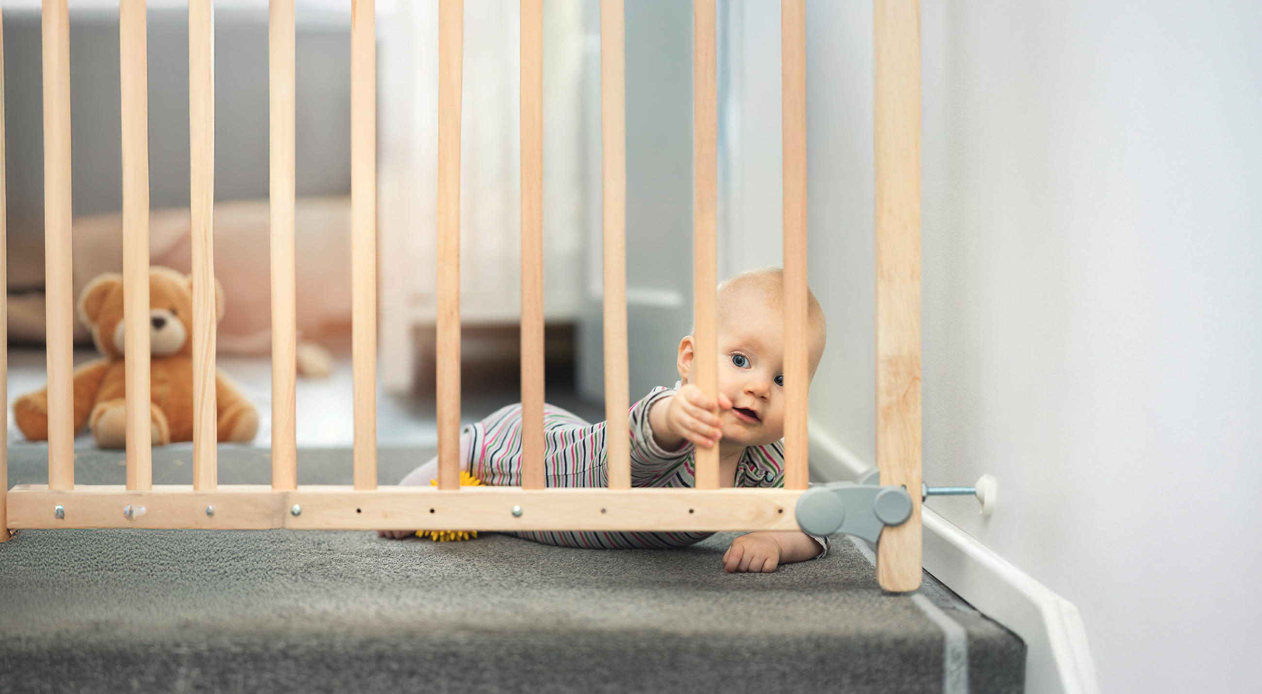 Ein Baby liegt auf dem Bauch vor einem Treppenschutzgitter und streckt die Hand neugierig durch die Holzstäbe. Es trägt einen gestreiften Einteiler, und im Hintergrund sitzt ein braunes Stofftier auf dem Teppich.