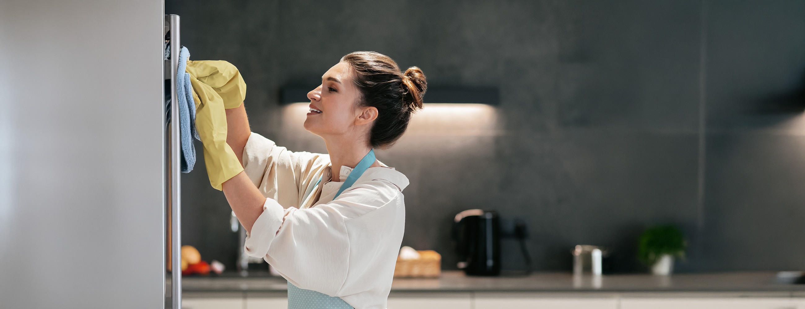 Frau steht in moderner Küche und wischt mit Handschuhen und Putzlappen einen silbernen Kühlschrank von außen.