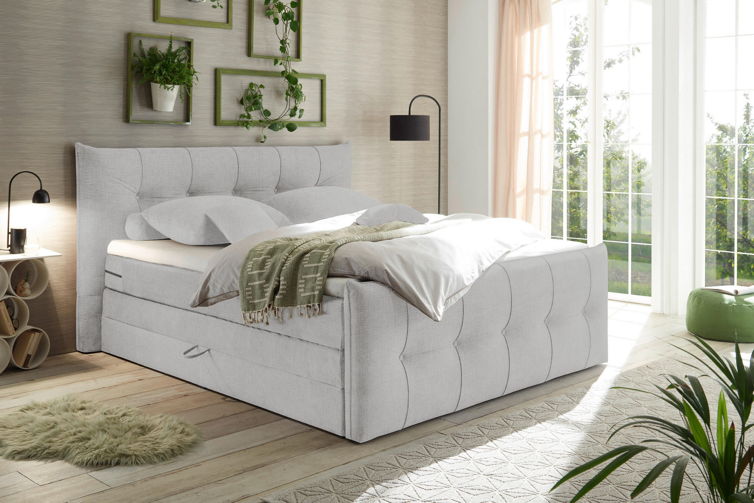  Ein modern eingerichtetes Schlafzimmer mit hellem Boxspringbett, weichen Textilien und großen Fenstern, durch die viel Tageslicht in den Raum fällt.