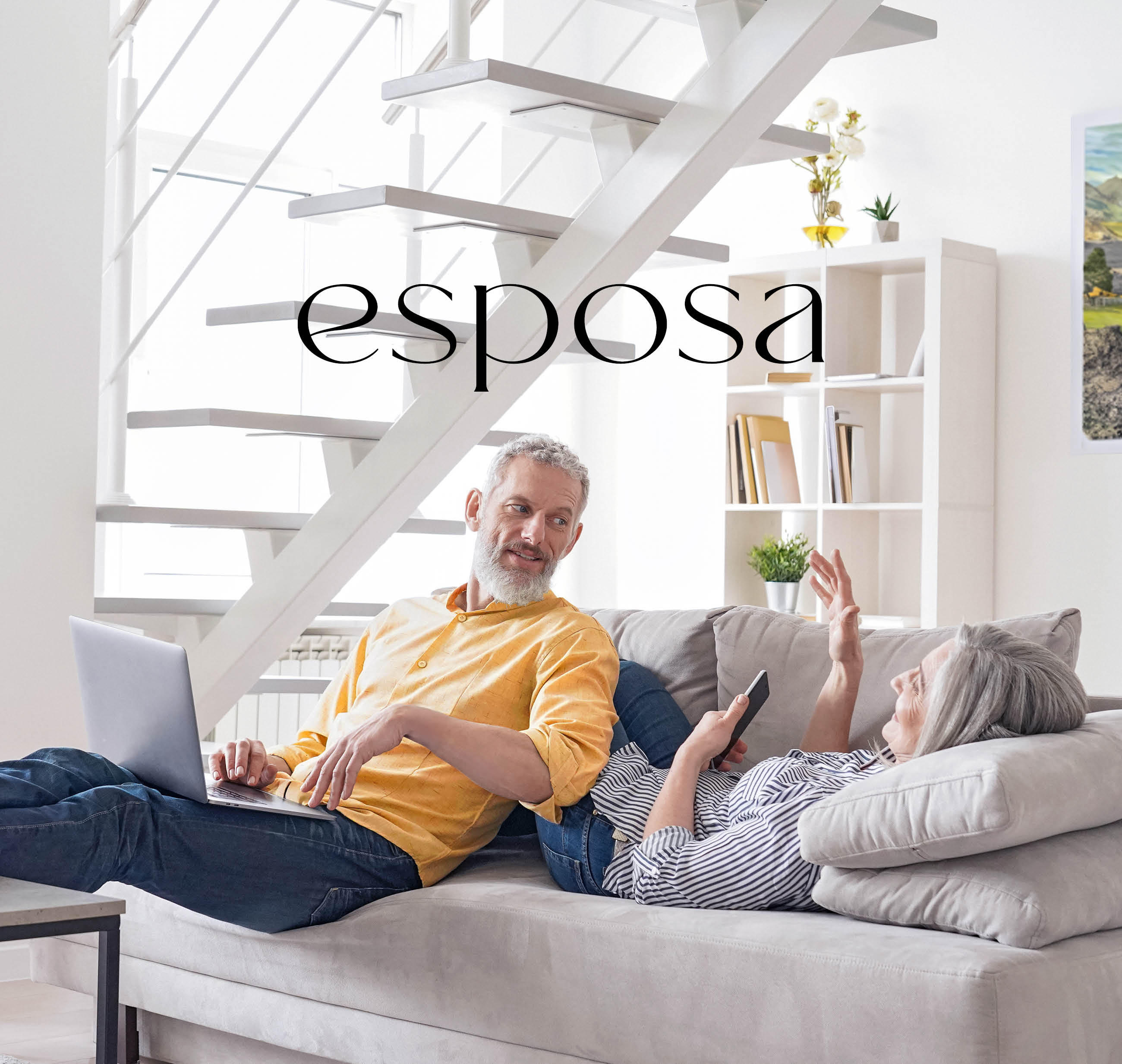 Zwei Personen entspannen auf einem modernen Sofa unter einer offenen Treppe, während sie miteinander sprechen und digitale Geräte nutzen.