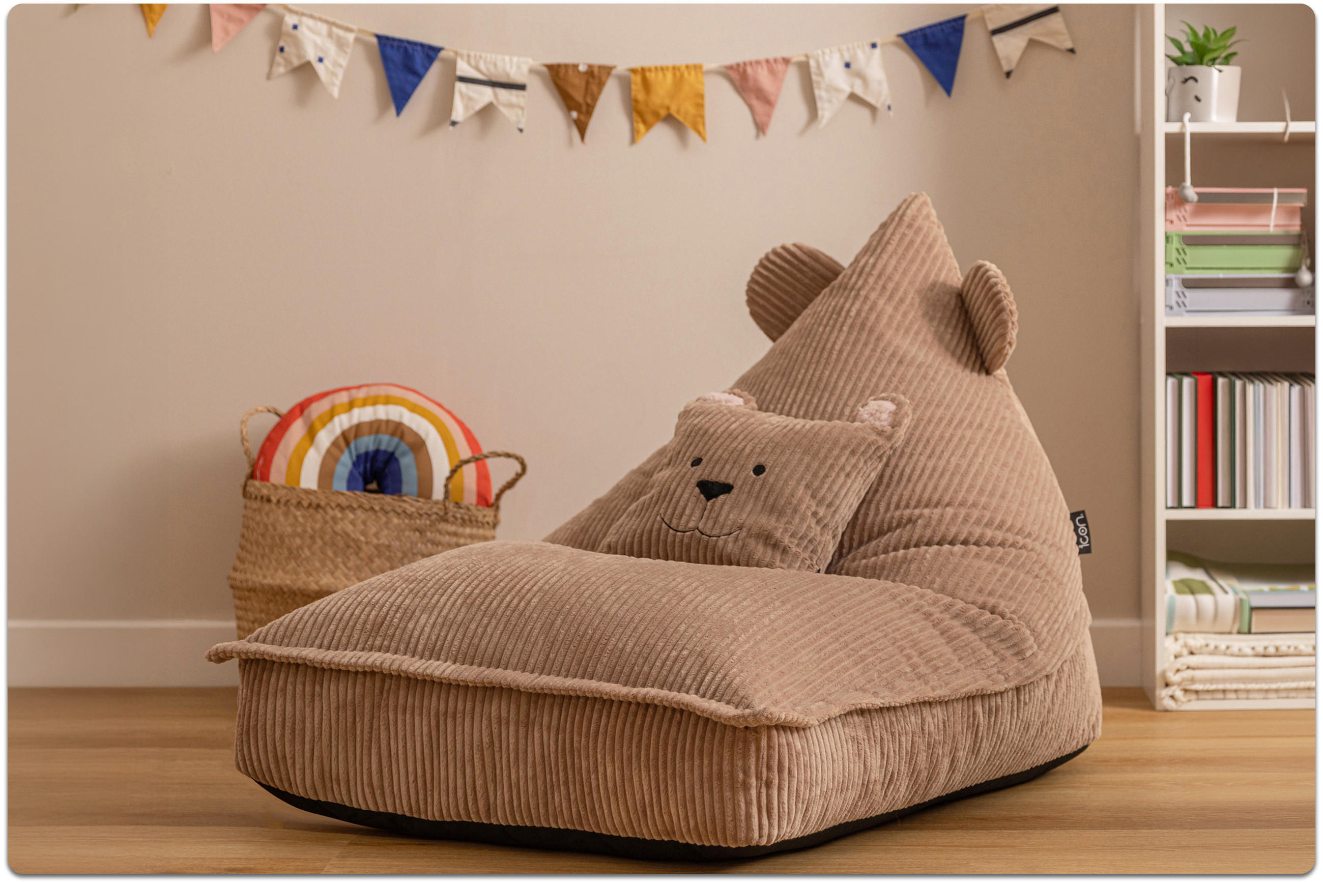 Beiger, cordförmiger Sitzsack mit Bärenohren und passendem Kissen in einem Kinderzimmer mit bunter Wimpelkette und Korb mit Regenbogenkissen.