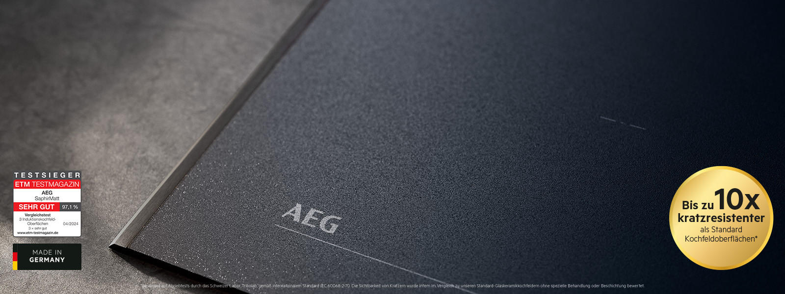 Nahaufnahme eines mattschwarzen AEG Induktionskochfelds mit integrierter Abzugseinheit; links eingeblendetes Testsieger-Siegel und „Made in Germany“, rechts Hinweis „Bis zu 10x kratzresistenter als Standard-Kochfeldoberflächen“.