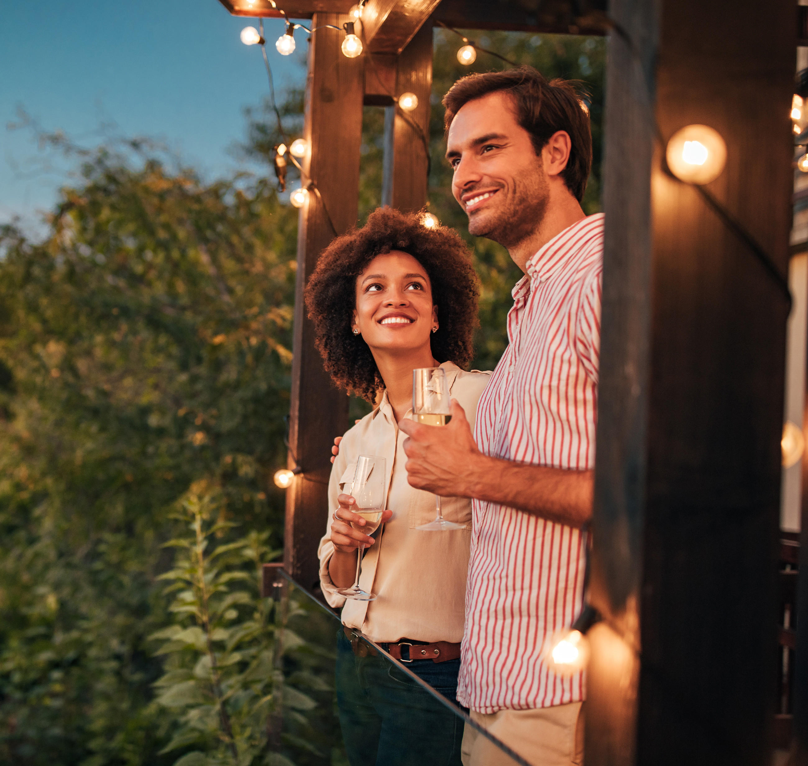 Ein Mann und eine Frau stehen auf einer Terrasse, umgeben von Lichtern, und genießen einen Sommerabend im Garten.