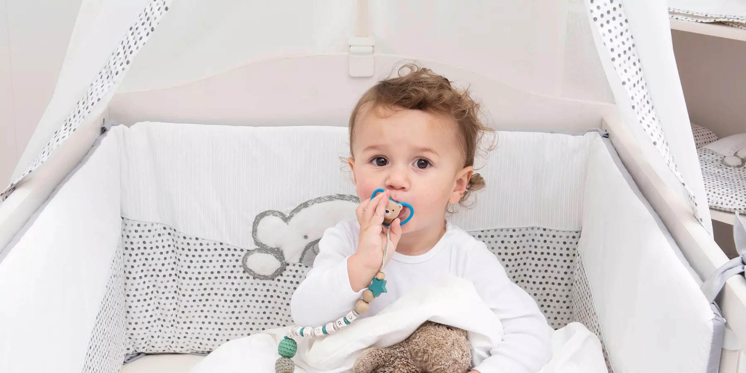 Baby im Gitterbett mit weiß-grauer Bettwäsche und Bärchen-Stickerei, hält Schnuller mit Perlenkette