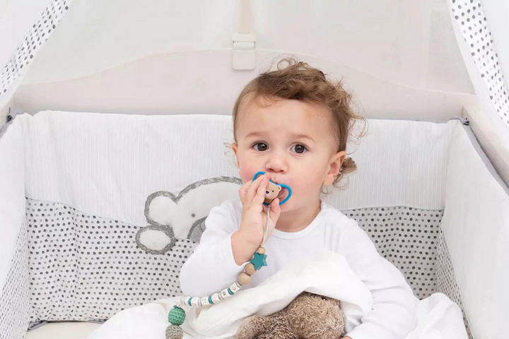 Baby im Gitterbett mit weiß-grauer Bettwäsche und Bärchen-Stickerei, hält Schnuller mit Perlenkette