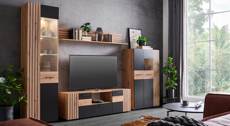 Wohnzimmer mit einer stilvollen Wohnwand aus Holz und schwarzen Elementen, bestehend aus Schränken, Regalen und einem TV. Dekoriert mit Pflanzen, Büchern und Accessoires vor einer grauen Wand