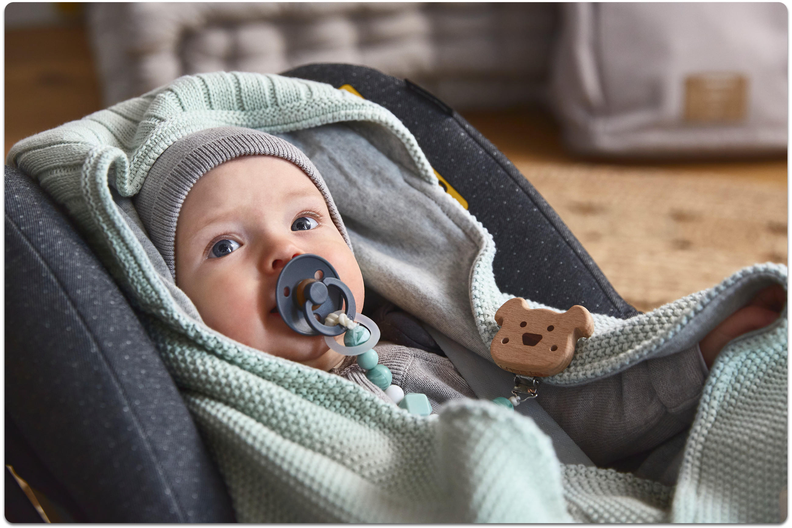 Baby liegt im dunkelgrauen Autositz, zugedeckt mit mintgrüner Strickdecke; sichtbarer Holz‑Schnullerhalter in Bärenform und pastellfarbene Beißkette.
