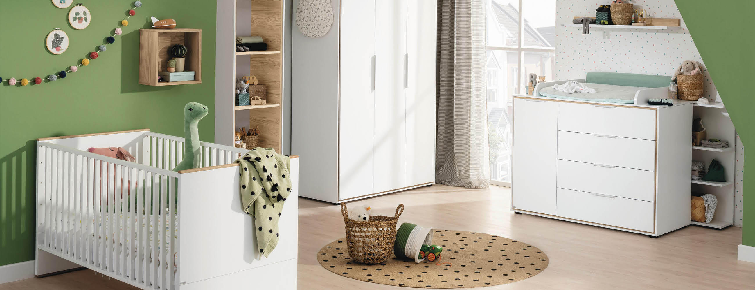 Kinderzimmer mit weißem Gitterbett, Kommode mit Wickelauflage, Spielzeugkorb und grüner Wanddekoration.