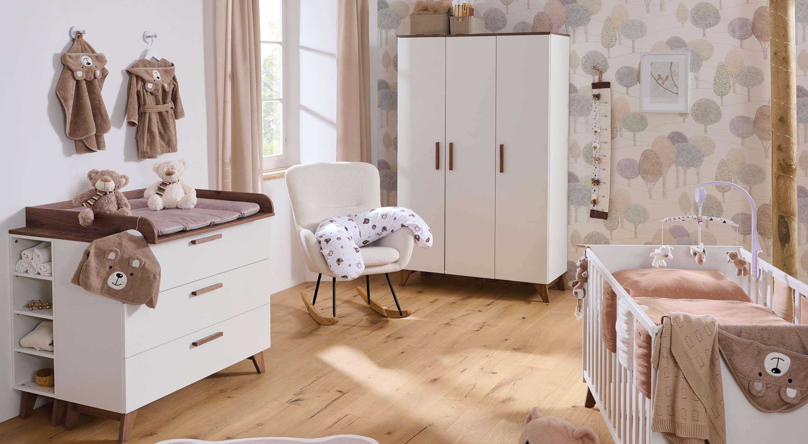 Helles Babyzimmer mit Wickelkommode und gepolsterter Auflage, Teddys und Bademänteln an der Wand, beigem Schrank, Schaukelstuhl mit Stillkissen sowie Babybett mit Mobile vor gemusterter Tapete.