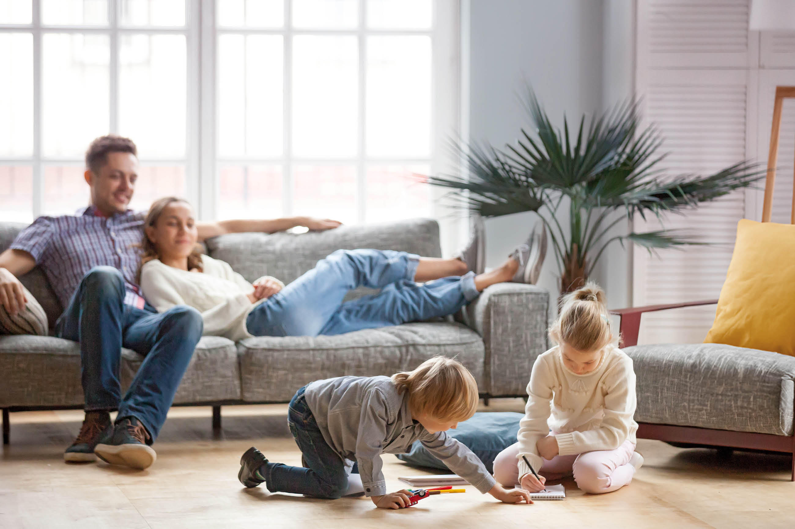 Zwei Erwachsene entspannen auf einem Sofa, während zwei Kinder davor auf dem Boden spielen und malen, wodurch eine wohnliche und ruhige Atmosphäre entsteht.